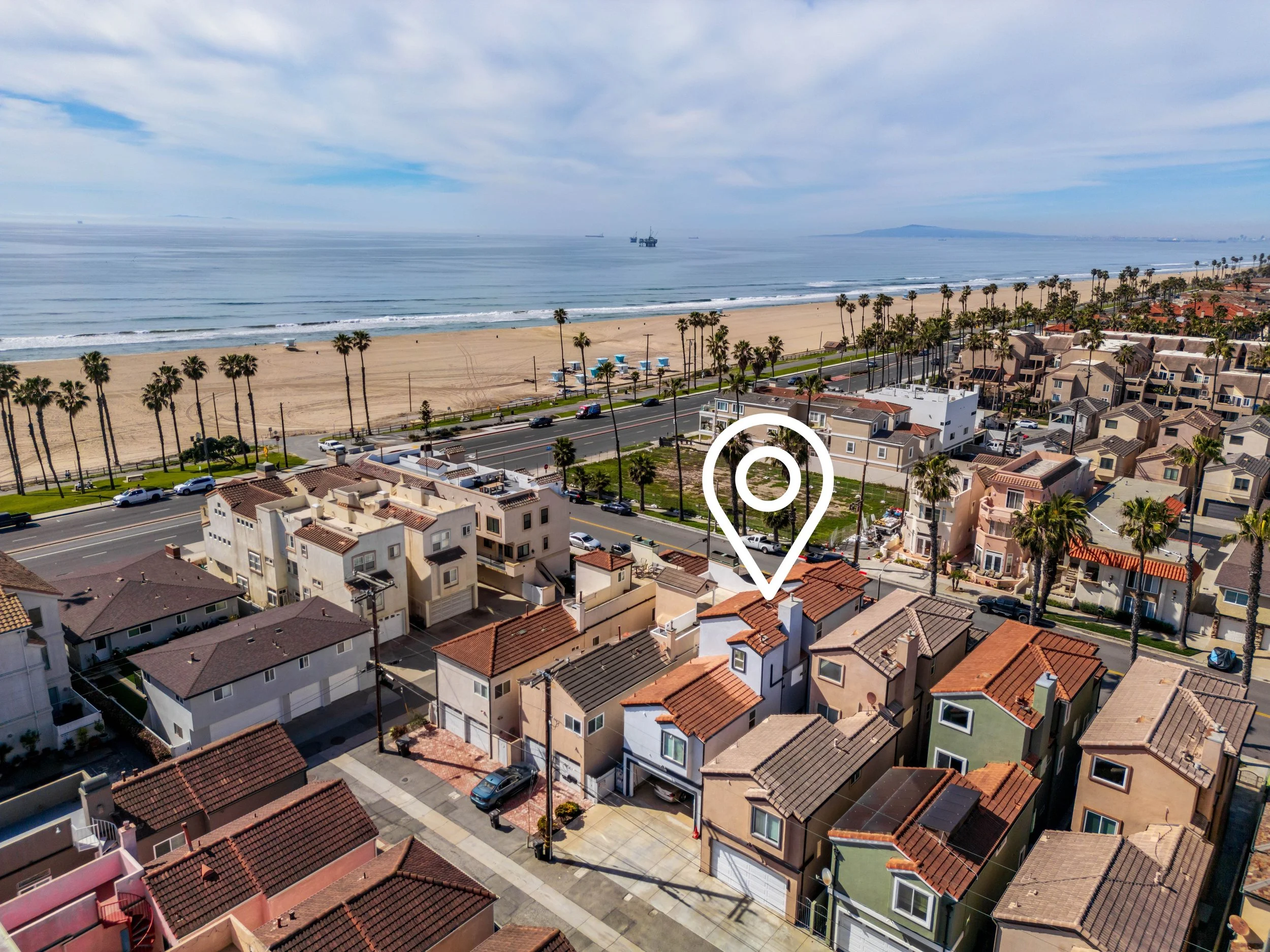 Aerial view of a neighborhood near a beach with houses, palm trees, roads, and parked cars, with the ocean and ships in the background under a partly cloudy sky.