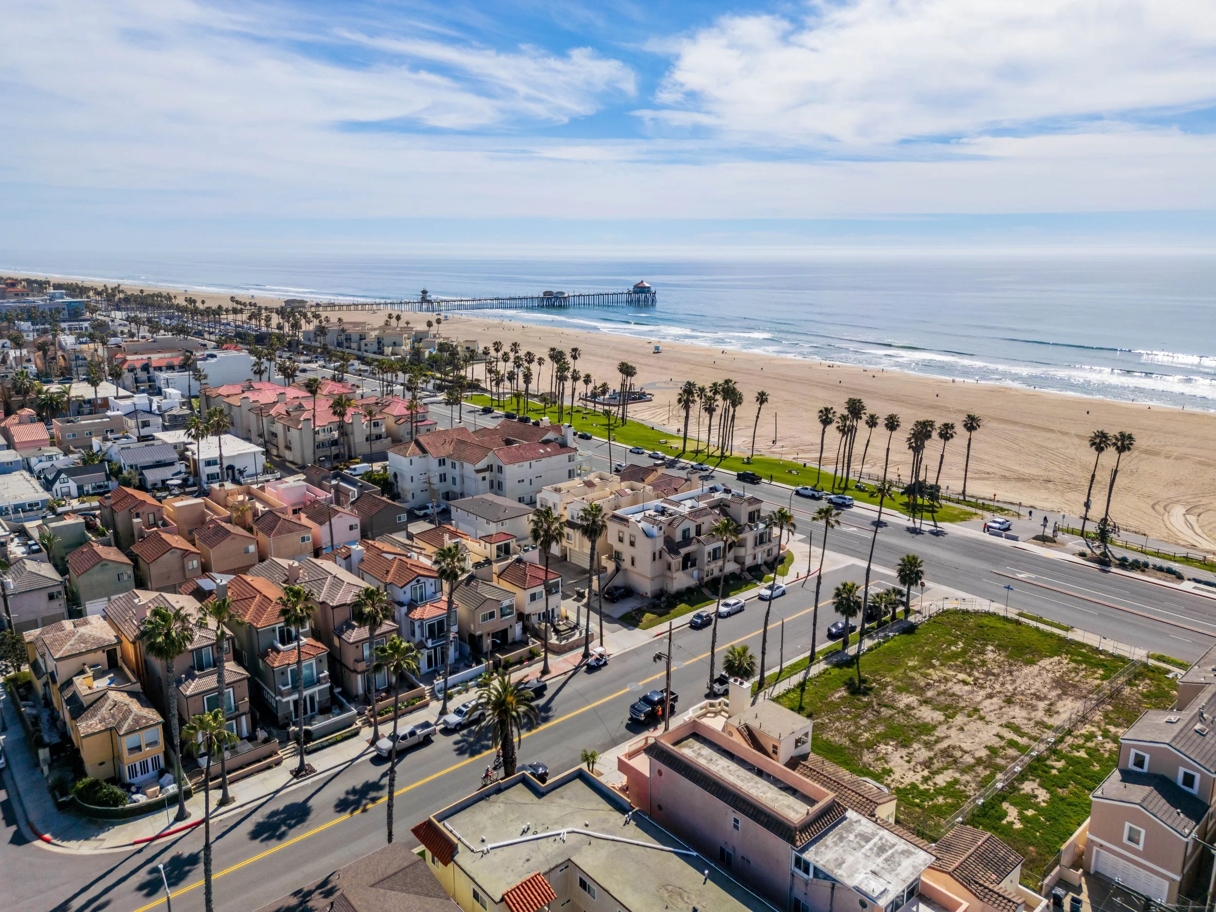 Aerial view of a beach city with houses, palm trees, a sandy beach, and a pier extending into the ocean under a partly cloudy sky.