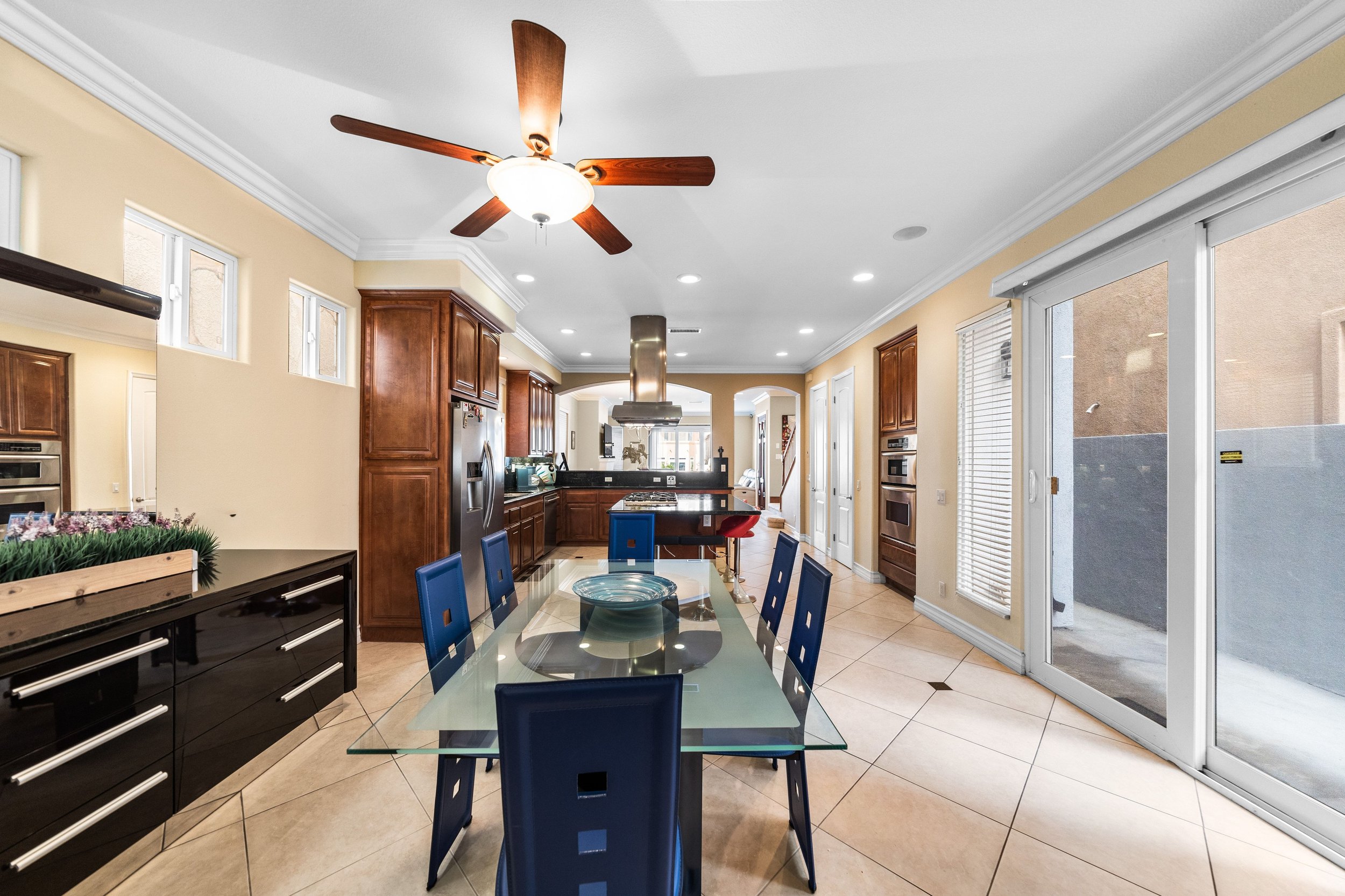 Kitchen with wooden cabinets, black stove, stainless steel refrigerator, glass dining table with blue chairs, ceiling fan, sliding glass door with blinds, and tile flooring.