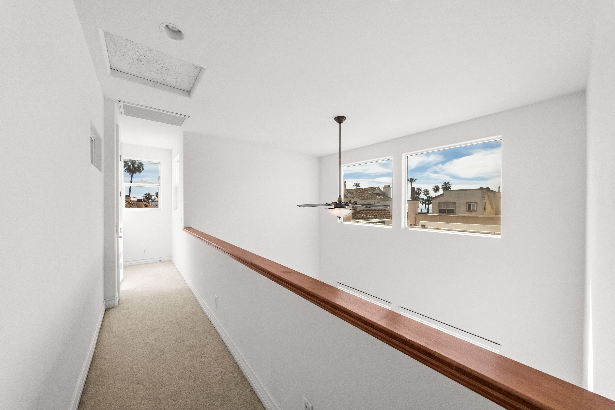 Interior view of a bright, white residential space with large windows showing palm trees and neighboring buildings, a ceiling fan, and a wooden railing.