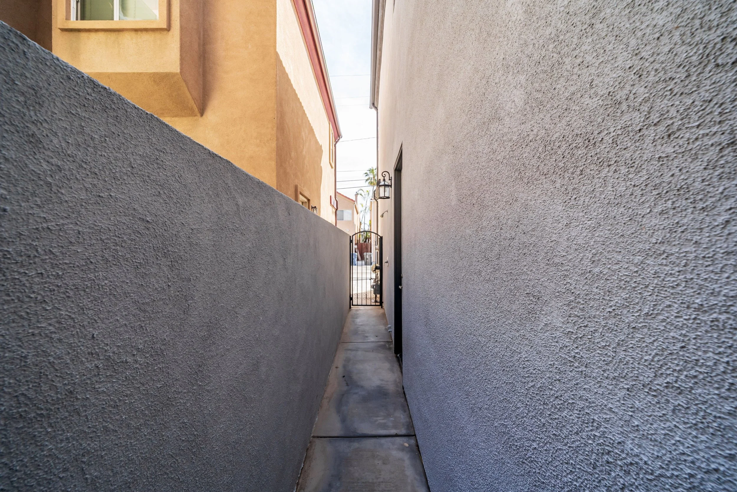 A narrow walkway between two textured exterior walls of a building, leading to a black gate at the back, with a lantern light fixture on the right wall and a building with a window and chimney visible beyond the gate.