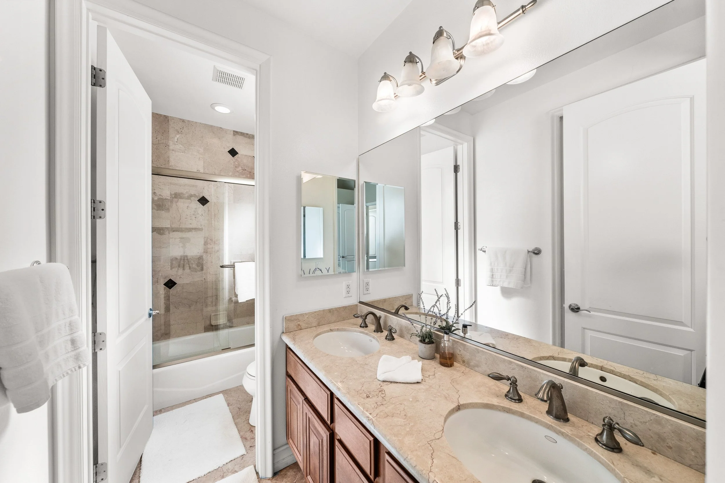 Bathroom with a large mirror, a single sink vanity with a granite countertop, and a glass-enclosed shower with beige and black tile. White walls and doors, with a towel hanging on the towel rack, and decorative plants on the countertop.