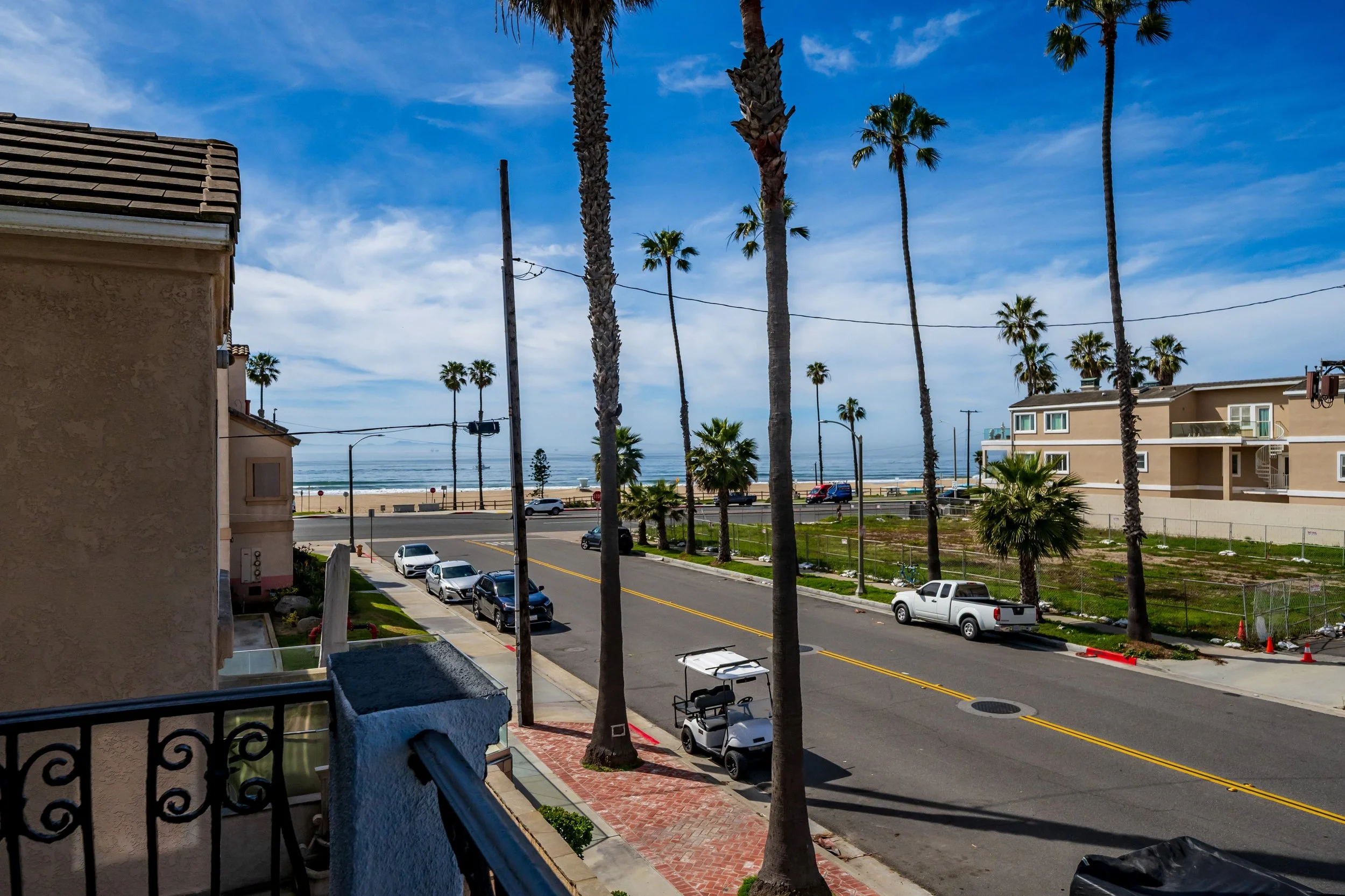 View from a balcony overlooking a street with parked cars, tall palm trees, and beach houses. In the background, the beach and ocean are visible under a blue sky with some clouds.
