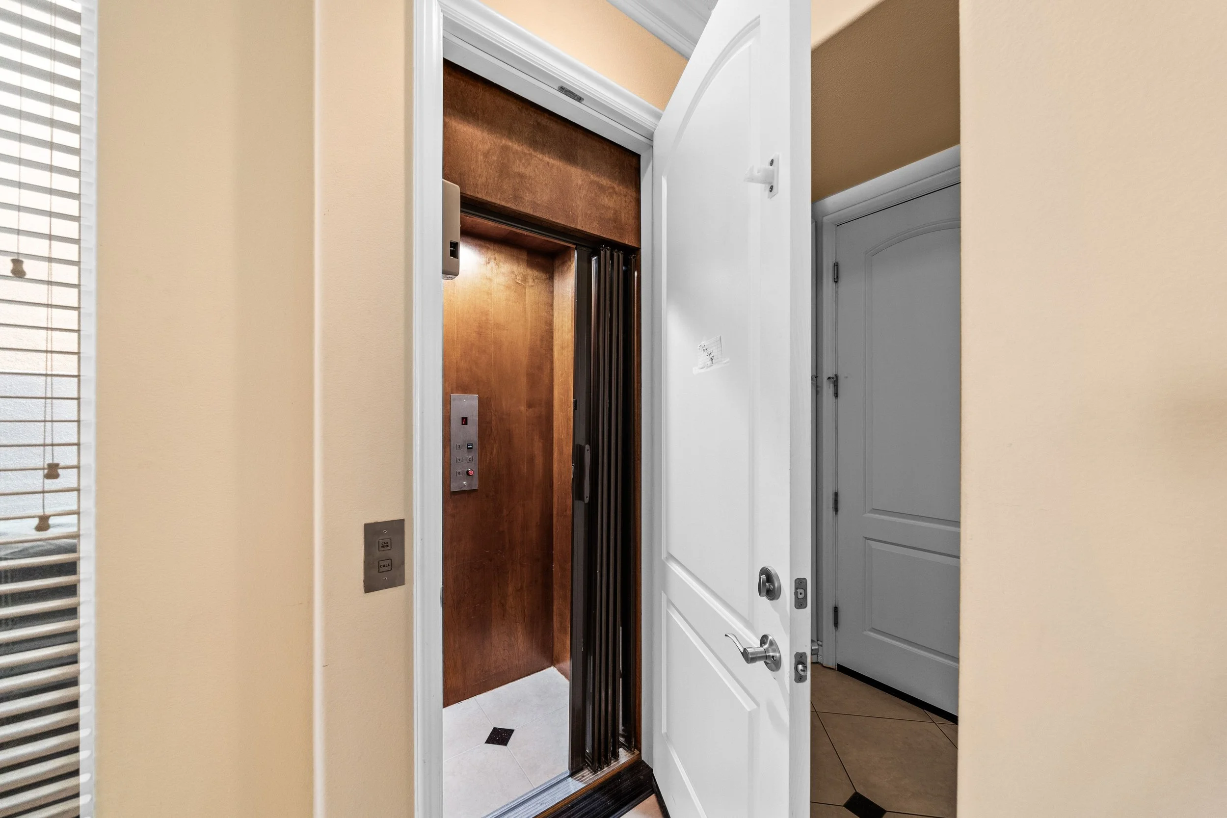 Open front door of an elevator showing a wooden interior with elevator controls, including buttons and a call button, on the wall outside the elevator.