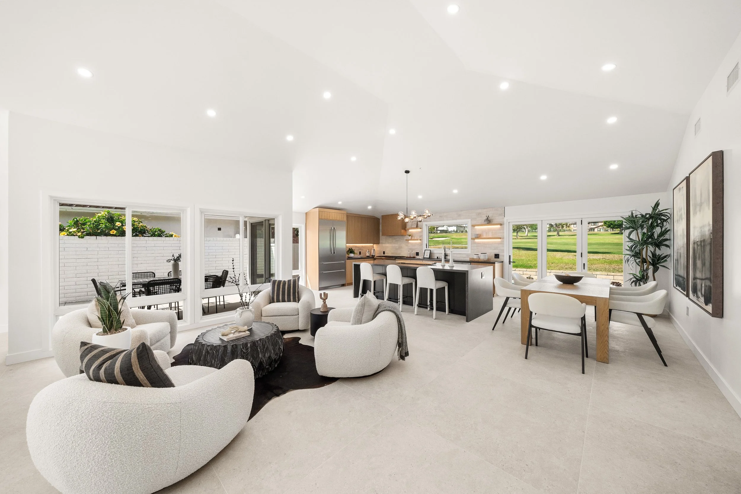 Open-concept living room, kitchen, and dining area with white walls, large windows, and modern furniture including white armchairs, a black and white sitting table, a dining table with white chairs, and a kitchen with wooden cabinets, black countertops, and bar stools. Large glass doors lead to an outdoor patio with a table and chairs, and green lawn with trees can be seen through the windows.