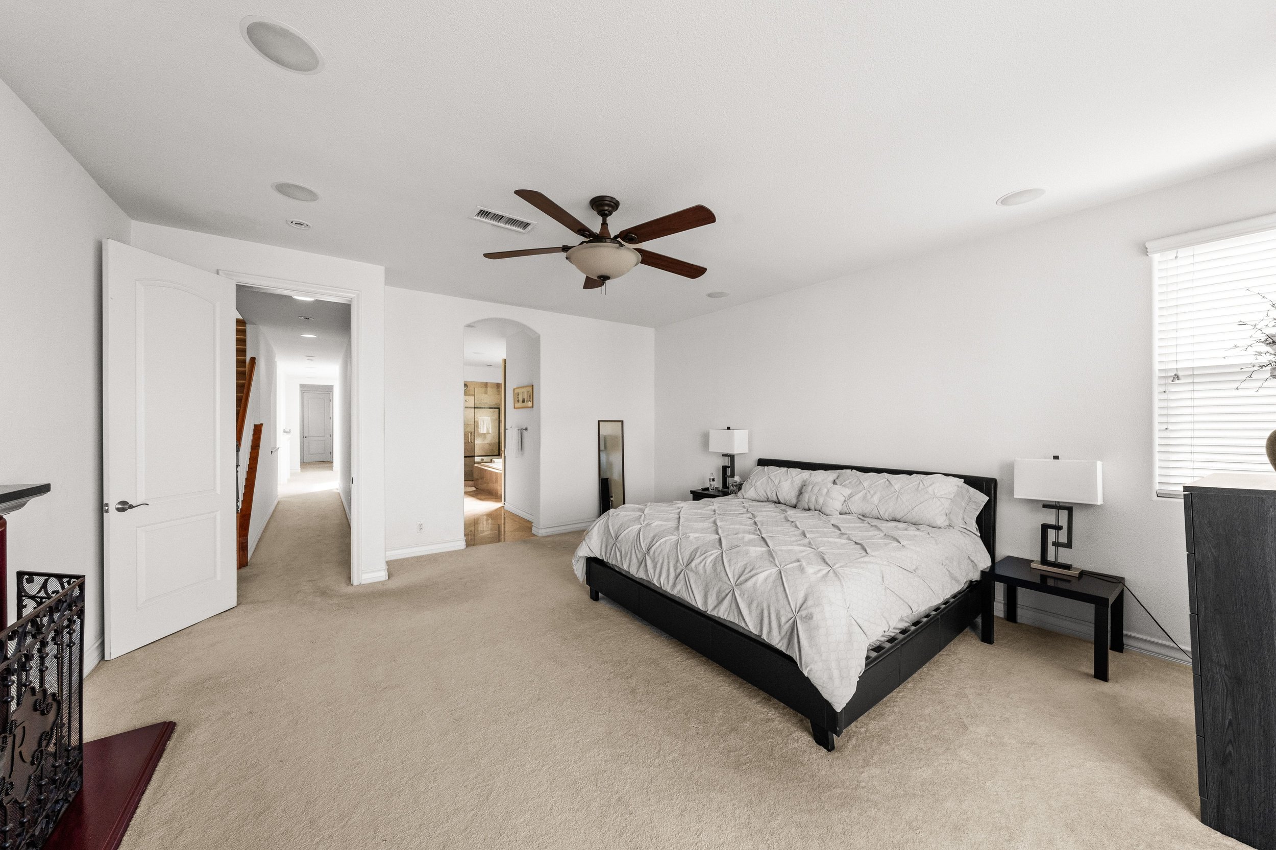 A spacious bedroom with white walls, a beige carpet, a black bed frame with a white quilt, and two black nightstands with white lamps. There is a ceiling fan with wooden blades and a light fixture. The room has a large window with white blinds, and a