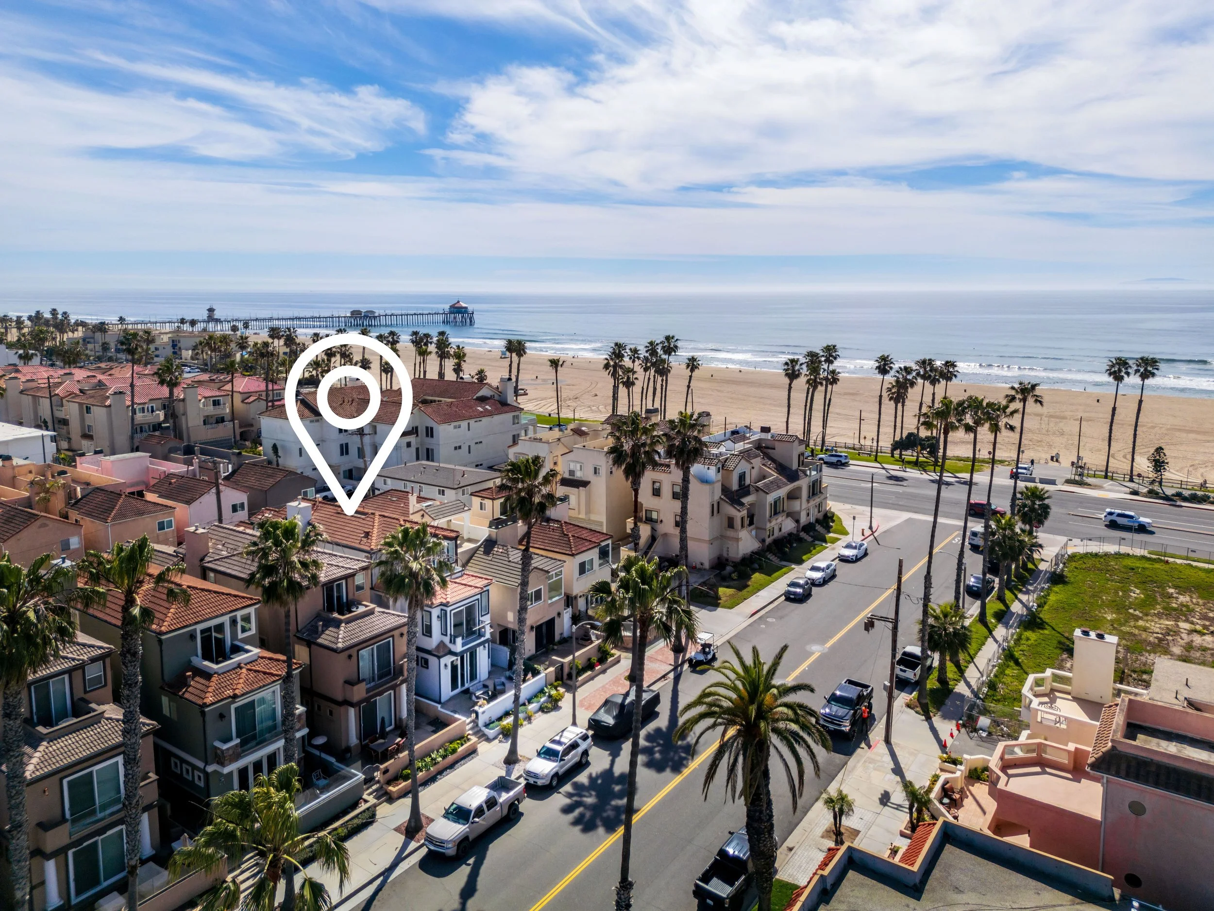 Aerial view of a beach town with residential buildings, palm trees, and a sandy beach leading to the ocean with a pier extending into the water and a blue sky with clouds.