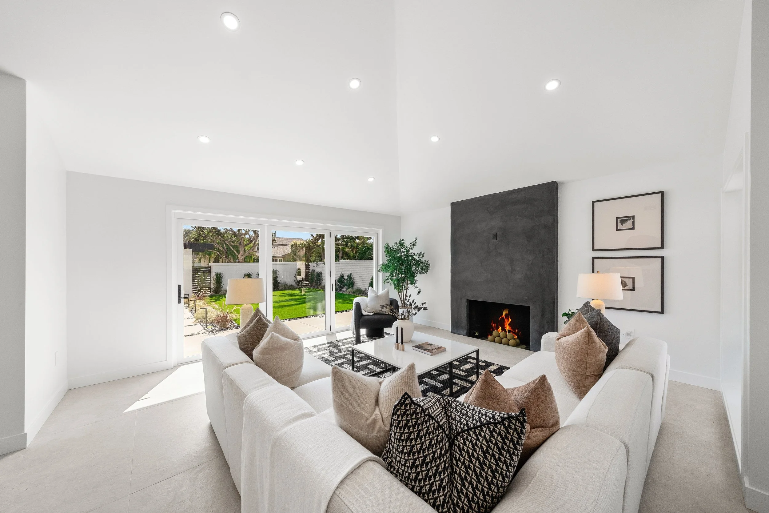 Living room with white sofas, beige pillows, a black armchair, a coffee table, a fireplace with a fire, framed artwork on the wall, and sliding glass doors leading to a backyard with green grass and trees.