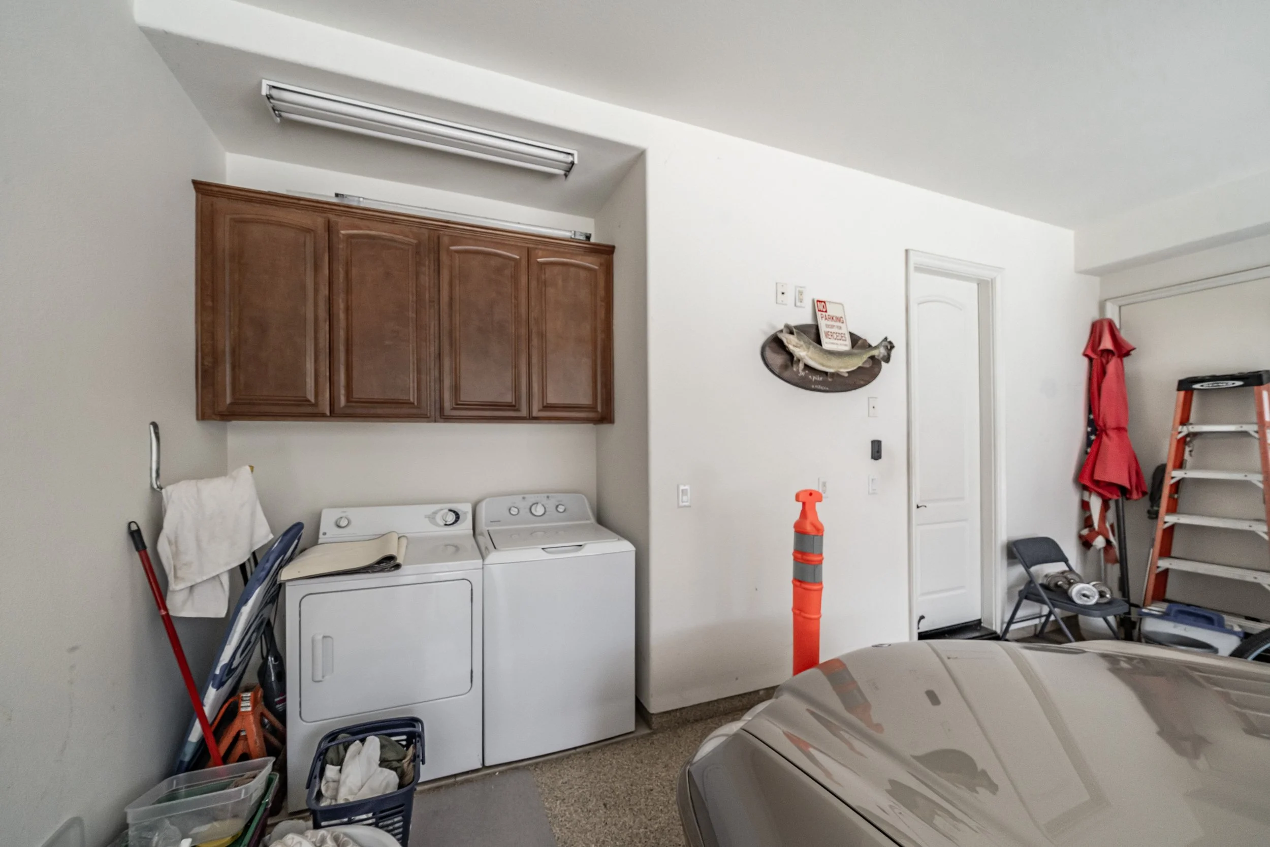 A laundry room with white walls and beige carpet, containing a washer and dryer, a wooden cabinet, various cleaning tools, storage bins, and a partially visible car in the foreground.