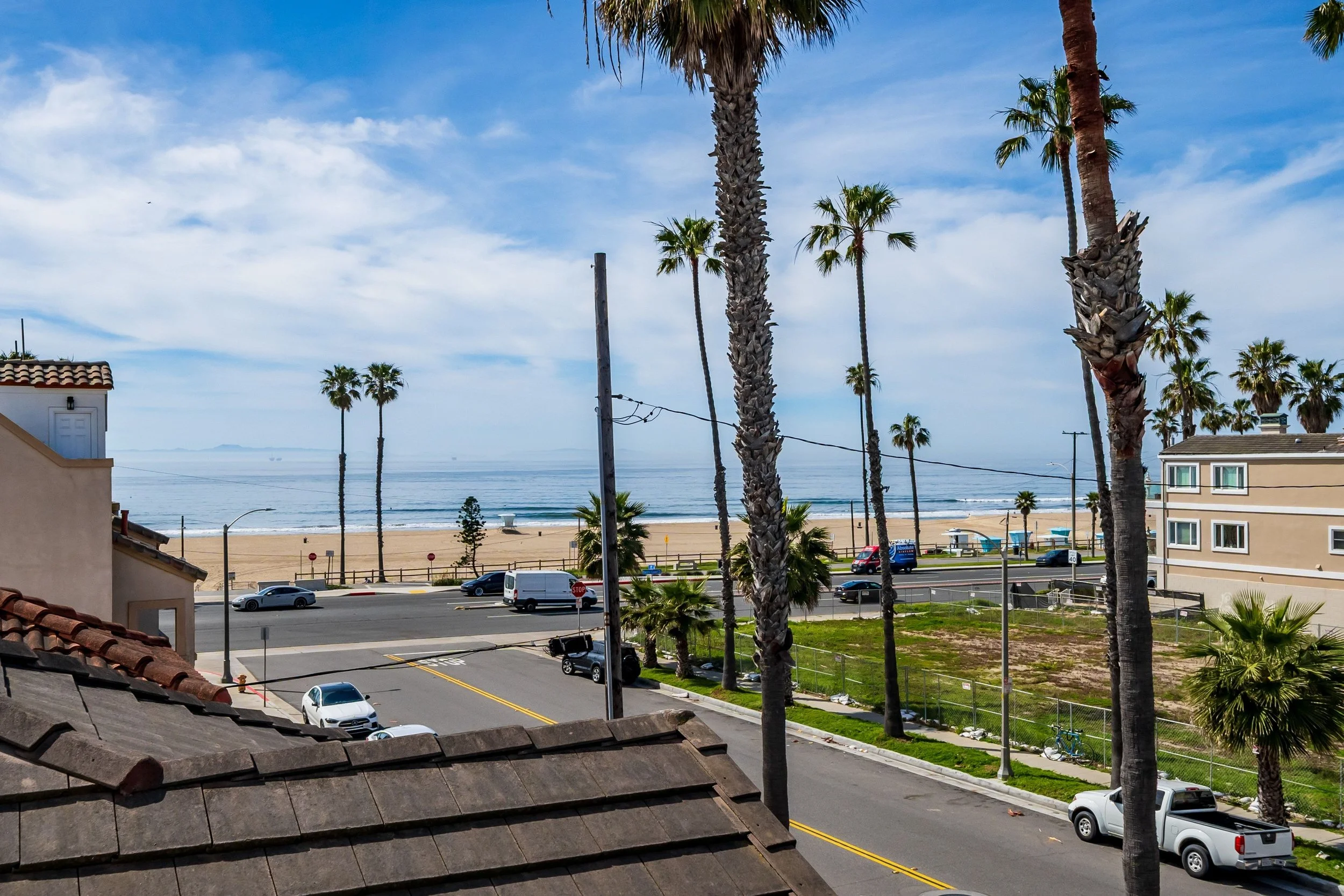 View of a coastal neighborhood with palm trees, ocean, sandy beach, houses, cars, and street, under a partly cloudy sky.