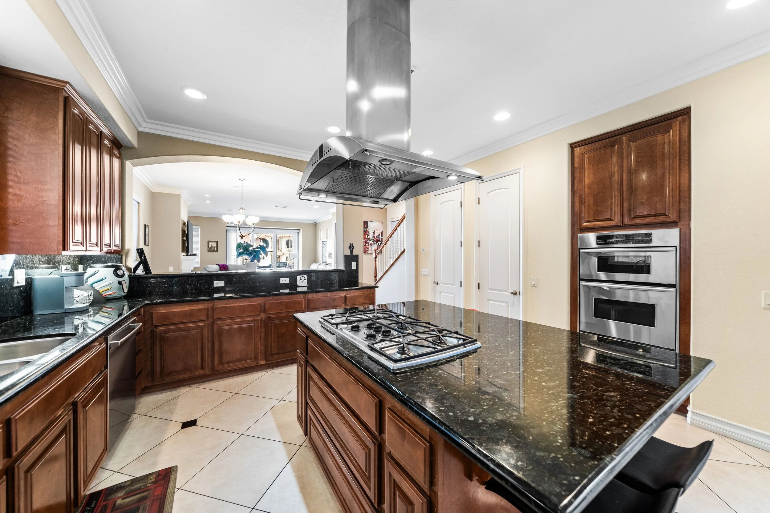 A spacious kitchen with wooden cabinets, black granite countertops, a gas stove with a range hood, built-in double ovens, and a dining area visible in the background.