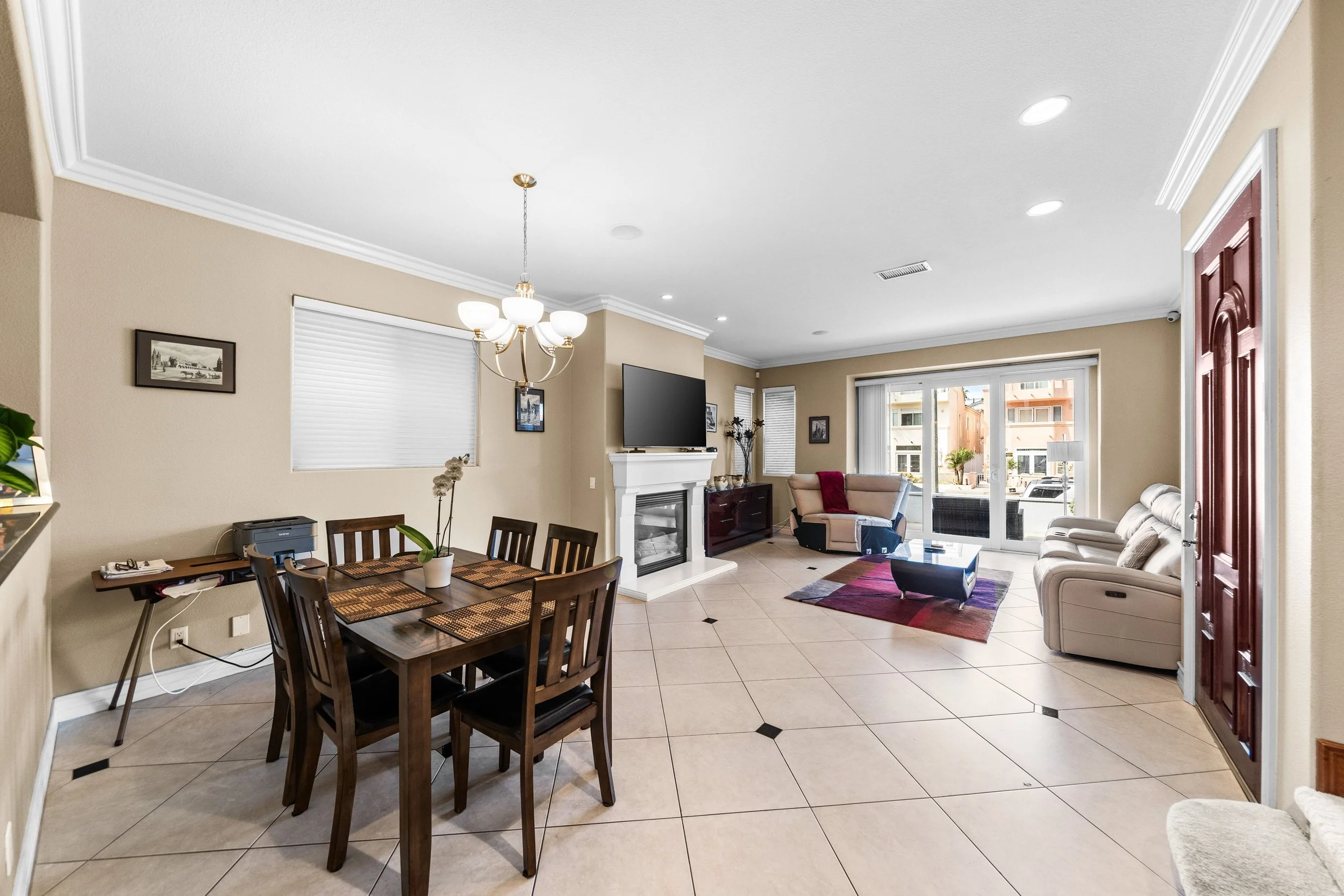 A spacious living room with a dining area, beige walls, tiled floor, a chandelier, a wall-mounted TV above a fireplace, a beige sofa, and large sliding glass doors leading outside.