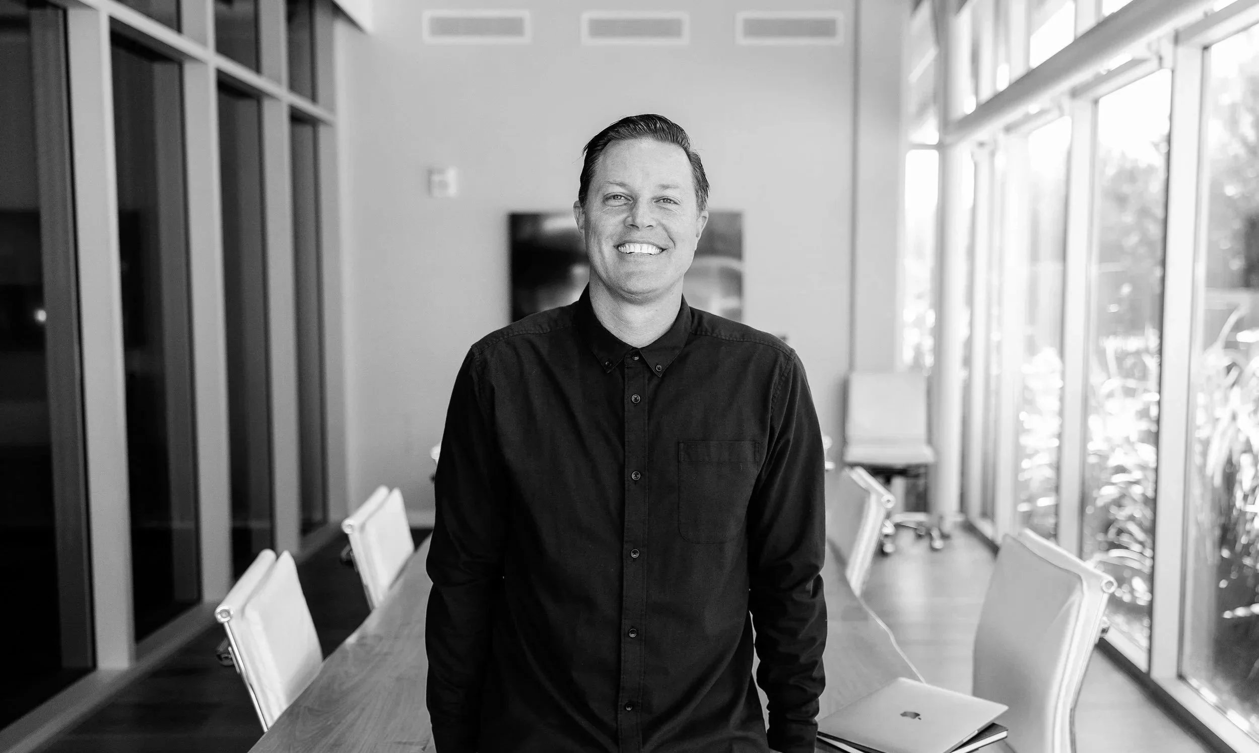 Black and white photo of a smiling man wearing a button-up shirt, standing in a modern office space with large windows along the right wall and chairs around a long table.