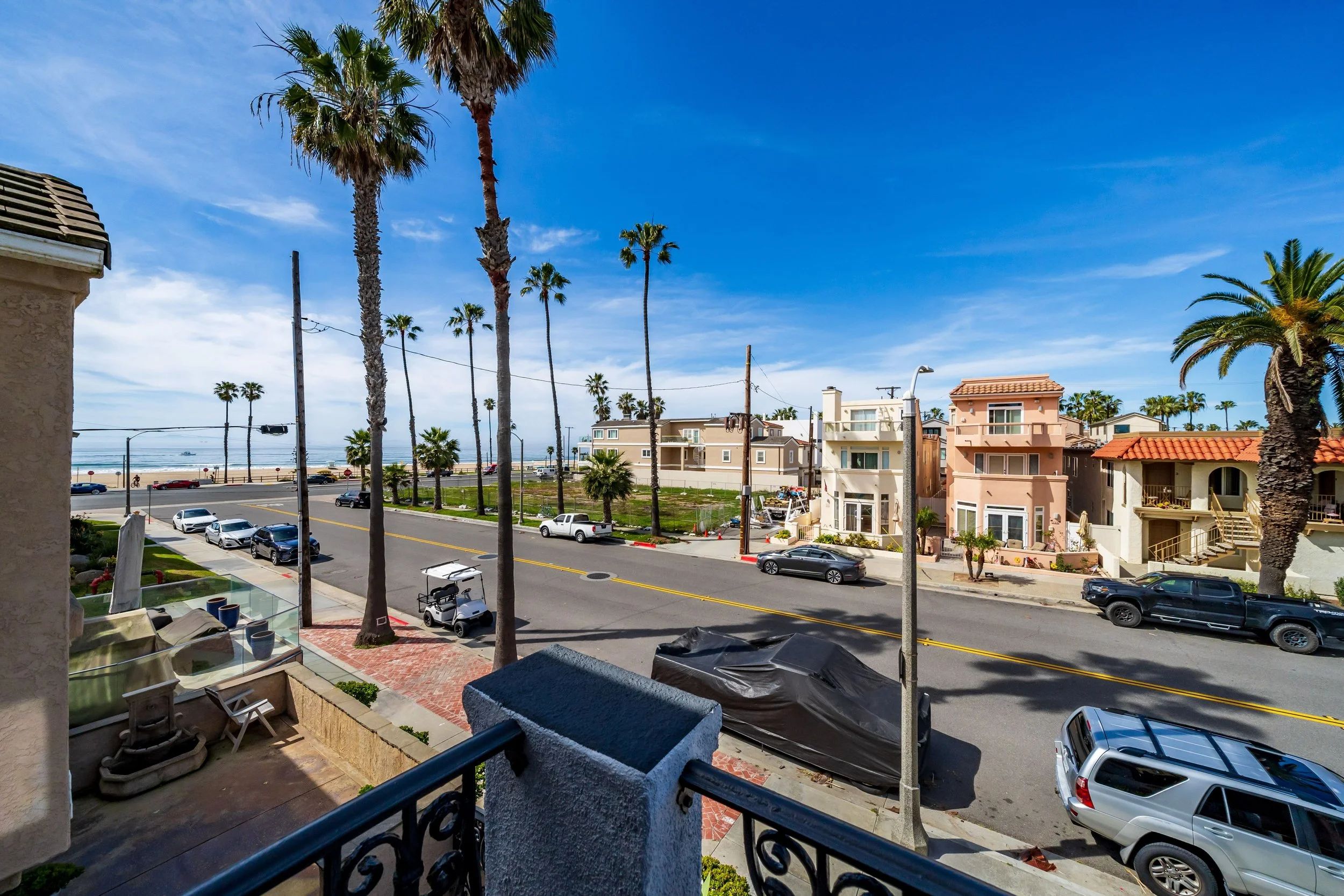 View from a balcony showing a street lined with palm trees, beach houses, and cars parked along the road, with the ocean and blue sky in the background.