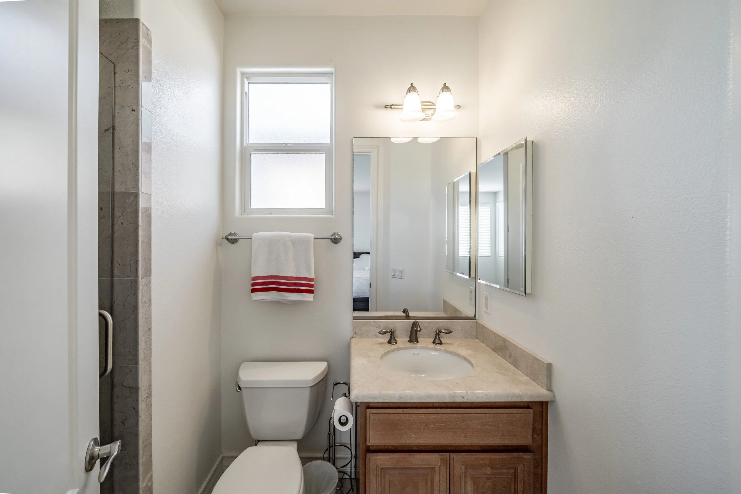 Small bathroom with a window above the toilet, a mirror, a beige vanity with a sink, a towel rack with a white towel with red stripes, a light fixture with two lights, and a shower area with a glass door and marble tiles.