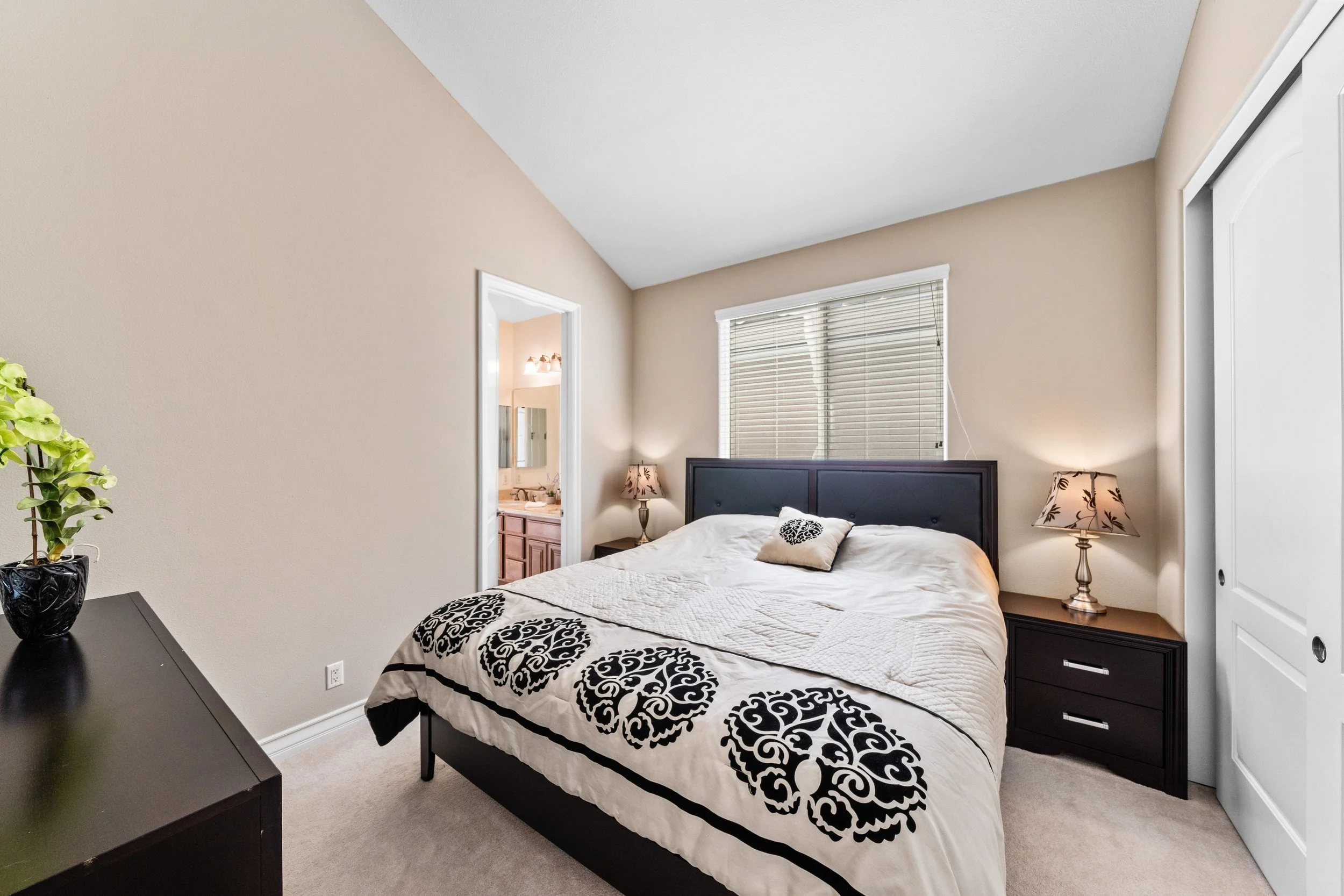 A bedroom with a black bed frame, black furniture, beige walls, and a window with blinds. There are lamps on nightstands on either side of the bed, and a mirror in the bathroom reflected in the doorway.