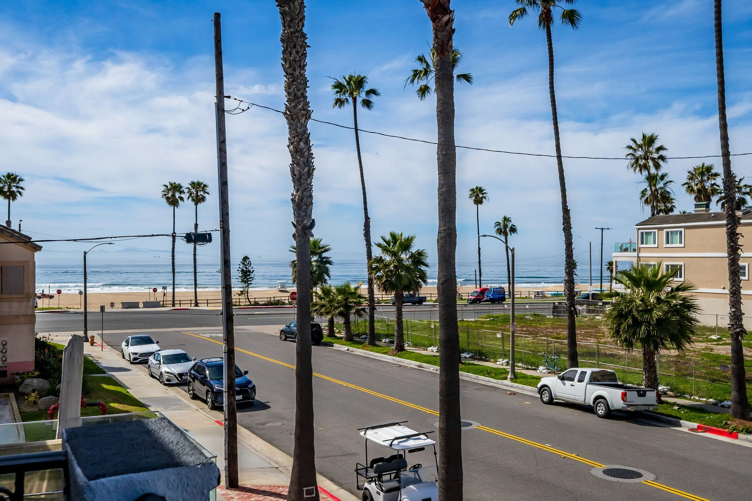 View of a beach with palm trees, a road with parked cars, houses, and the ocean in the background under a partly cloudy sky.