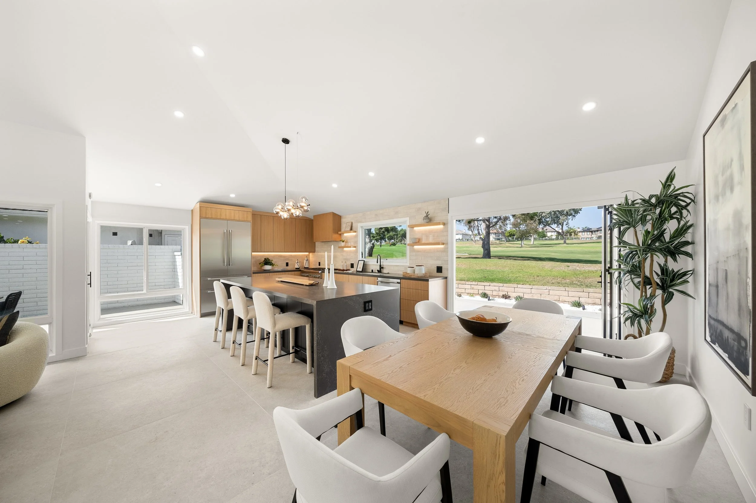 Open-concept kitchen and dining area with large windows view of a backyard with grass and trees, light-colored walls and furniture, wooden cabinets, modern lighting fixtures, and a kitchen island with barstools.