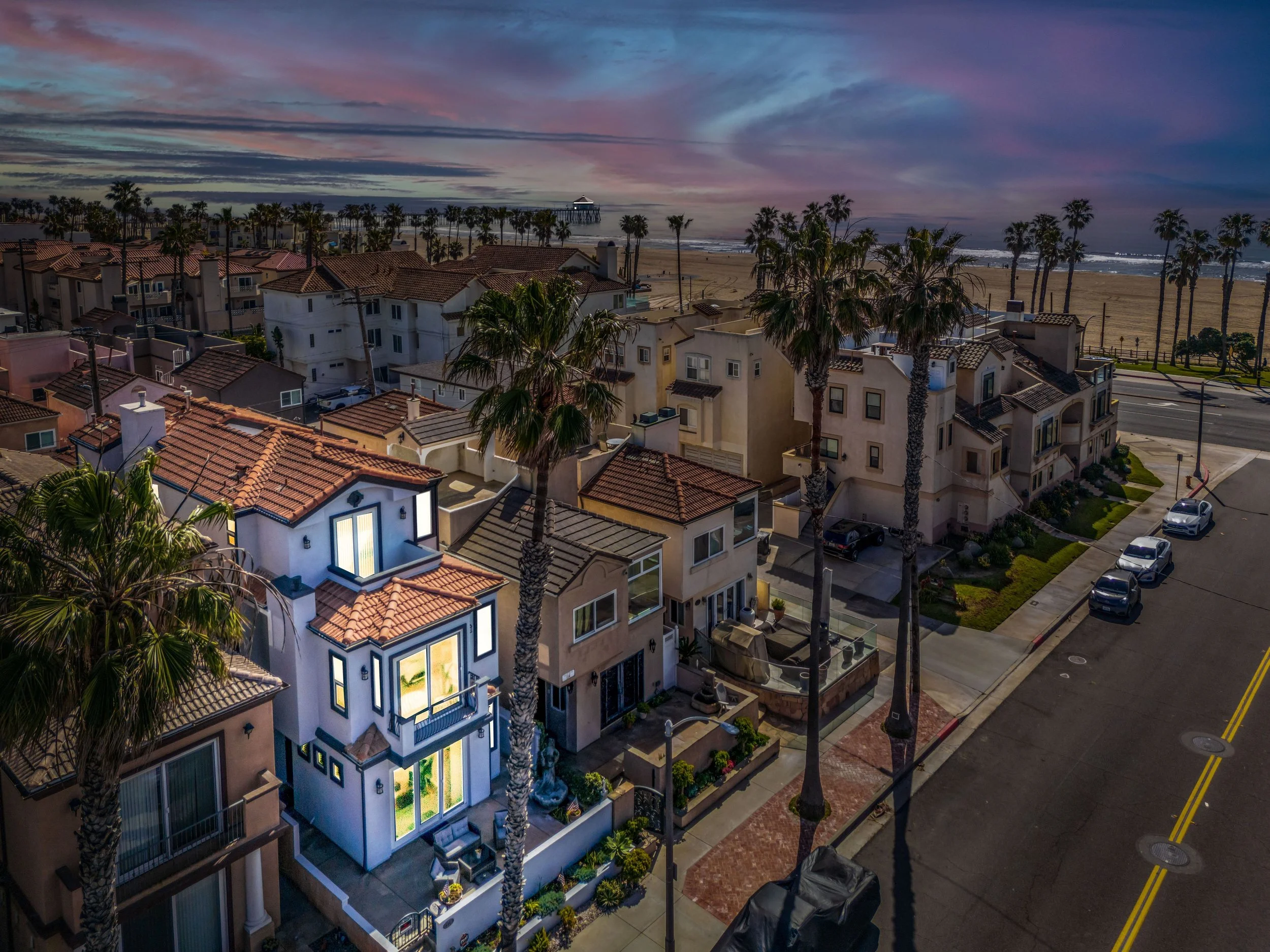 A view of a coastal neighborhood at dusk with houses, tall palm trees, and a sandy beach leading to the ocean with a pier in the distance. The sky is colorful with hints of pink, purple, and blue.