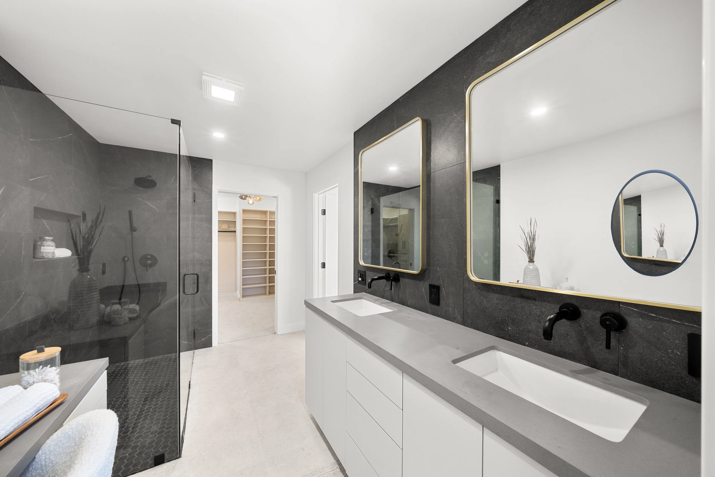 Modern bathroom with a vanity, double mirrors framed in gold, black fixtures, gray counters, and a walk-in shower with black tiles, gold accents, and a rainfall showerhead. A vase with decorative branches is on the counter, and another on a shelf with towels.