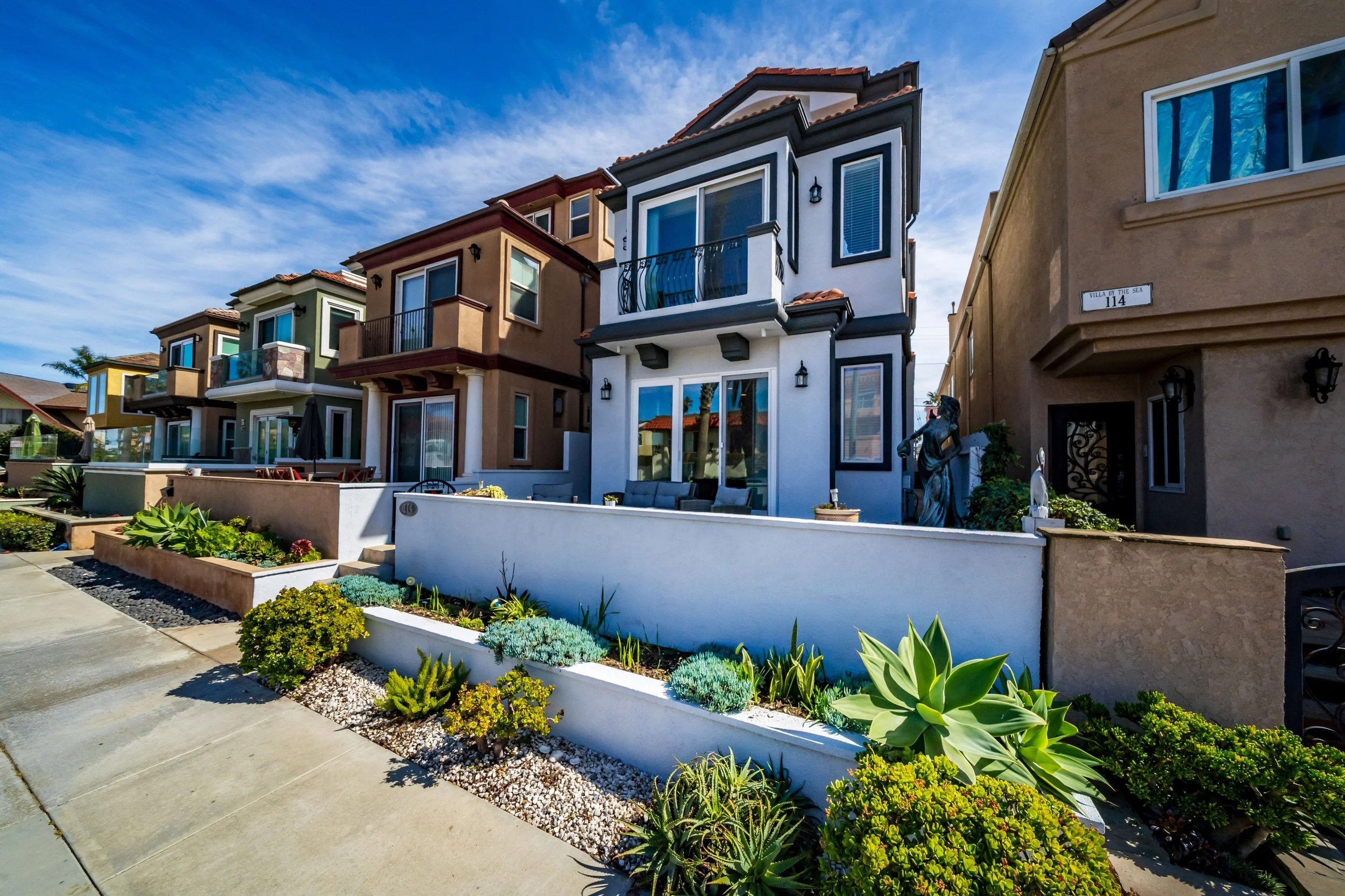 Colorful multi-story residential buildings with front gardens, a sidewalk, and a blue sky with wispy clouds.