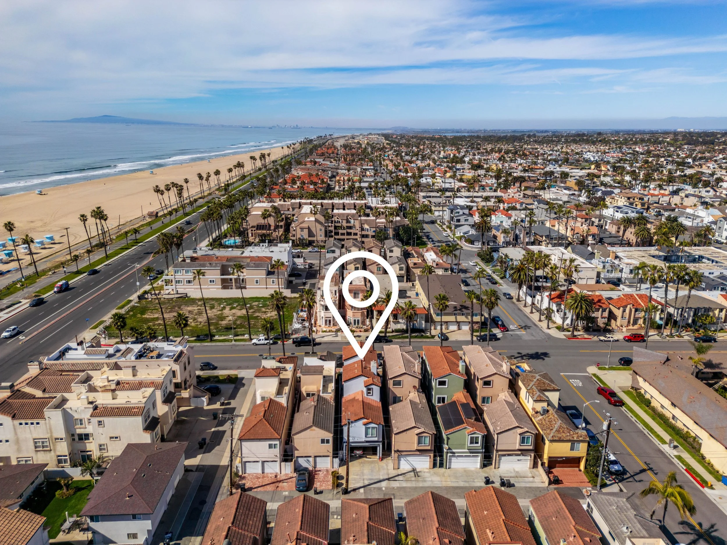 Aerial view of a coastal neighborhood with houses, palm trees, a street, beach, ocean, and mountains in the background.