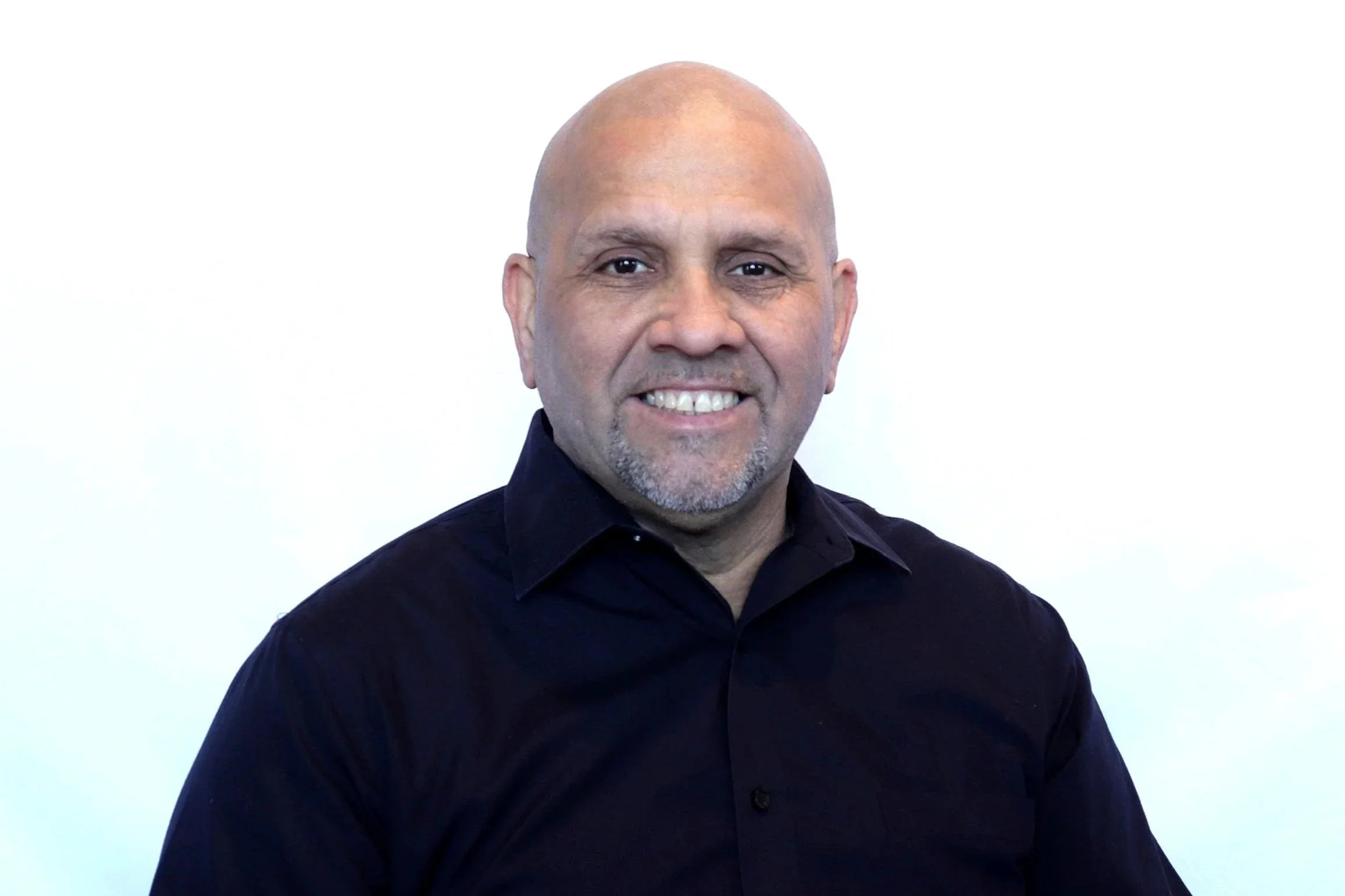 A middle-aged man with a shaved head and a goatee, smiling and wearing a black button-up shirt, posed against a plain white background.