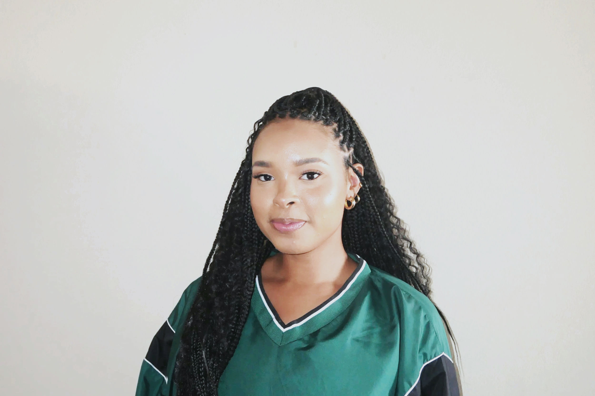 A young woman with long, braided hair wearing a green sports jersey and gold hoop earrings, standing against a plain light-colored wall.