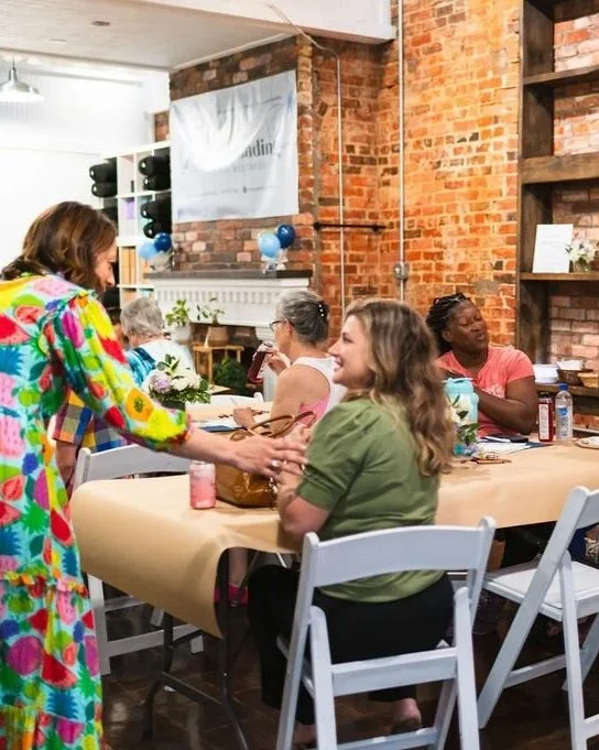 A woman in a multicolored floral dress is sitting at a table, getting her nails painted by another woman. The table is in a cozy, rustic indoor setting with brick walls and shelves, and other people are sitting and chatting in the background.
