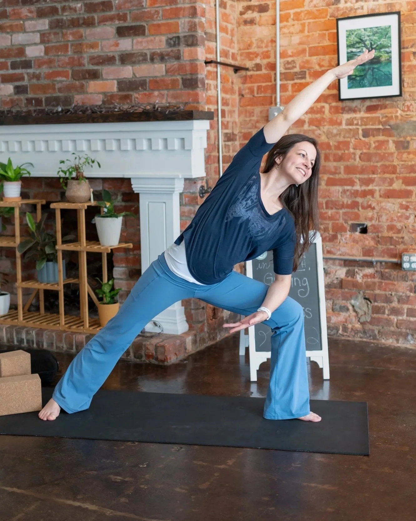 A woman practicing yoga indoors on a black mat, wearing a dark blue top and light blue pants, in a side lunge pose with one arm extended upward and the other downward.