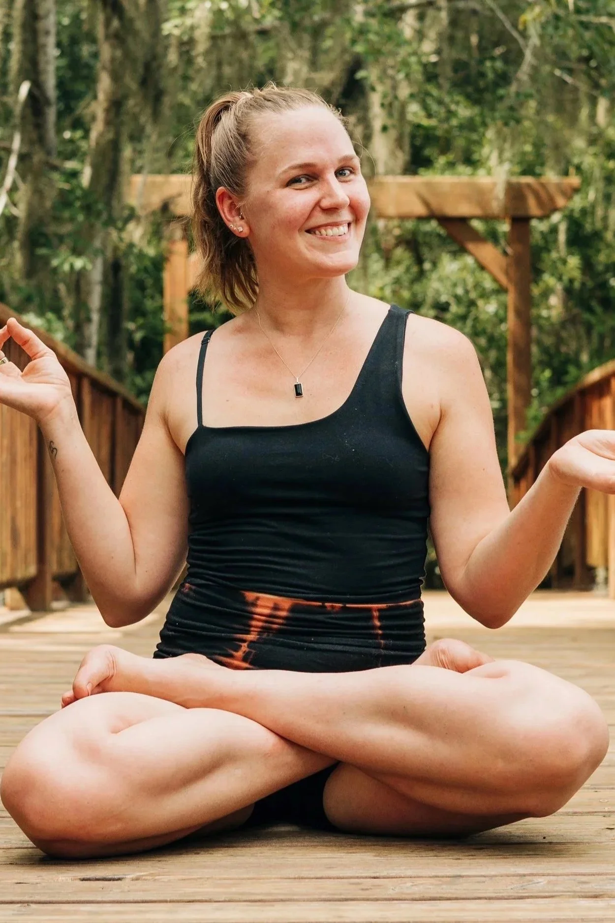 A woman practicing yoga outdoors on a wooden deck surrounded by trees, sitting cross-legged in a meditative pose with a smile on her face.