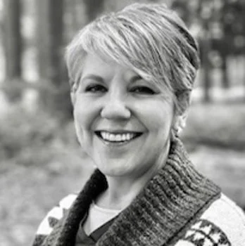 Close-up black-and-white photo of smiling woman with short hair, outdoors in a wooded area.