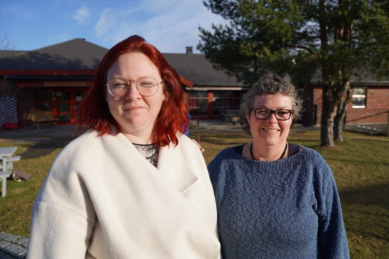 To smilende kvinner står ute i sollys foran Fontenehuset Rygge, iført briller og varme klær, i et rolig uteområde.