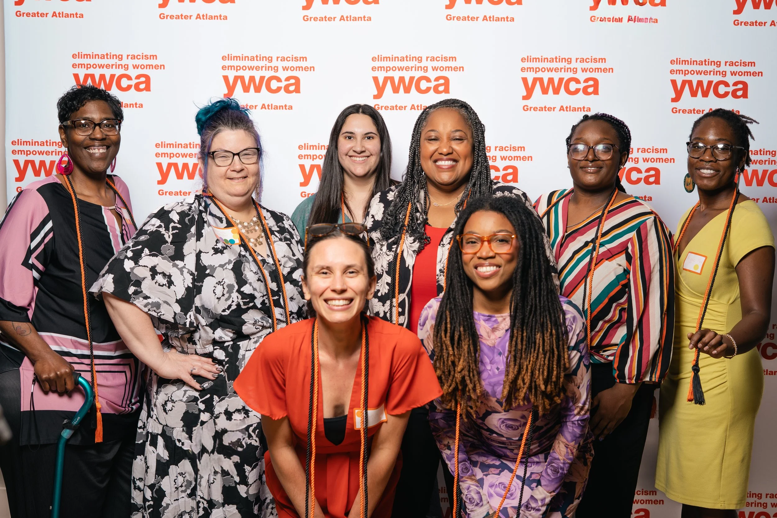 Six women standing together inside a building, smiling at the camera, holding a document, with wood-paneled walls and columns in the background.