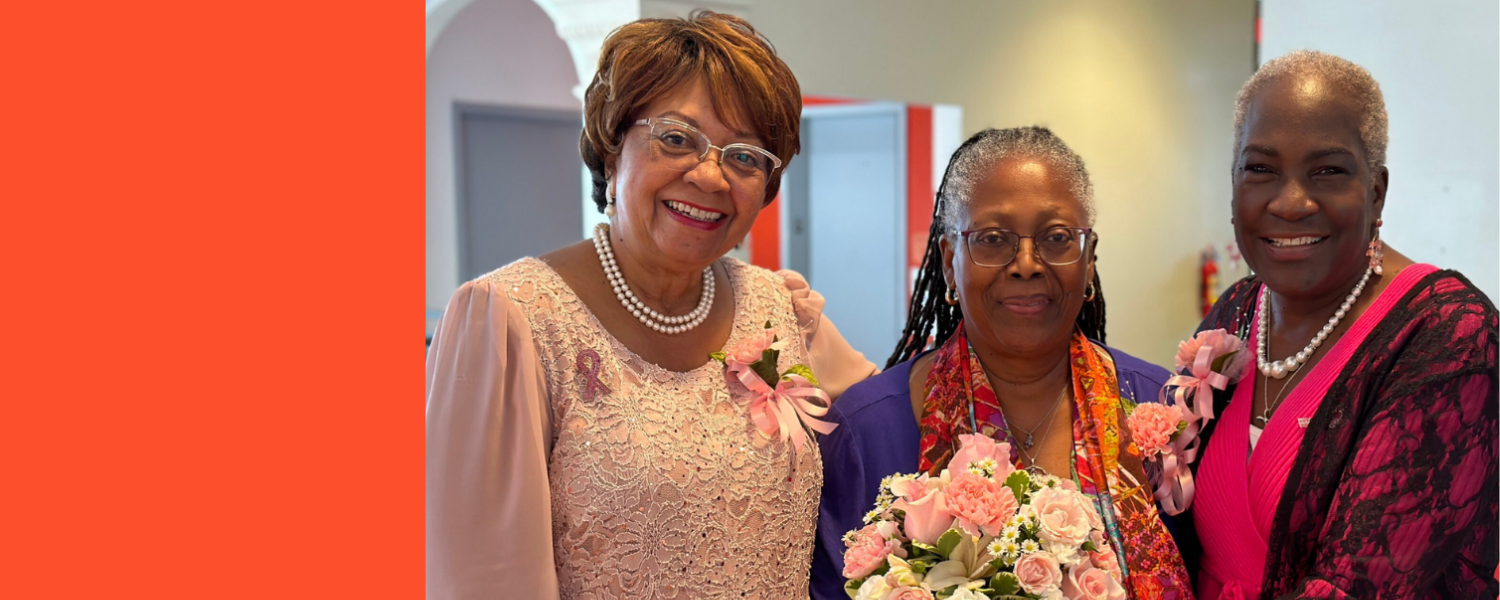 Three women smiling and posing together at a celebration, with the woman in the middle holding a bouquet of pink and white flowers. They are dressed in colorful attire with floral corsages and jewelry, standing indoors.