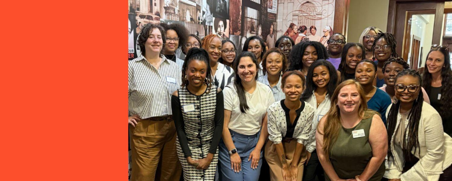 Group of diverse people standing on steps outside a historic building, smiling at the camera, some wearing backpacks and colorful clothing.