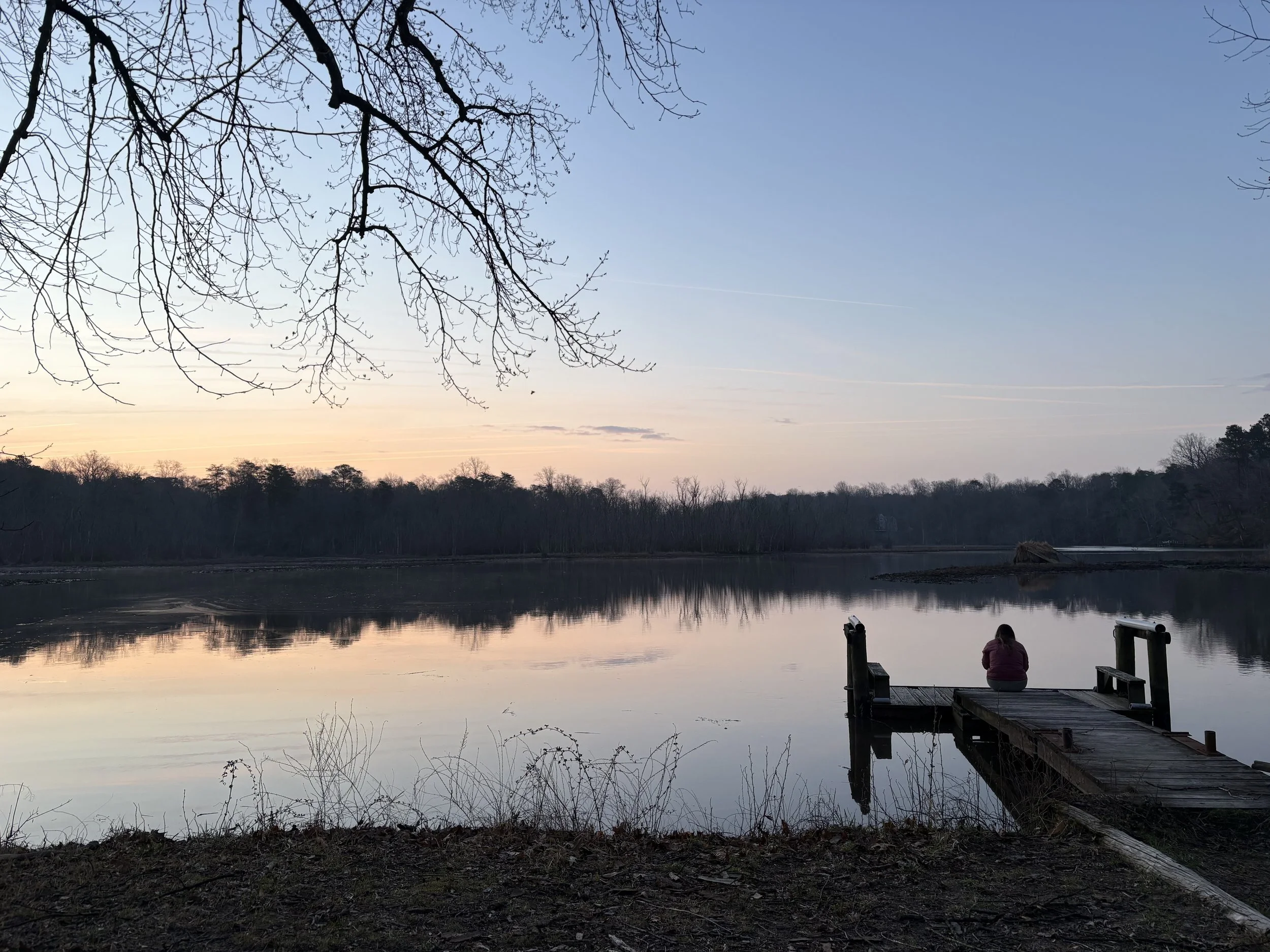 At sunrise on the edge of a calm creek with woodland in the distance, a woman sits with her back toward the viewer as she gazes across the water.