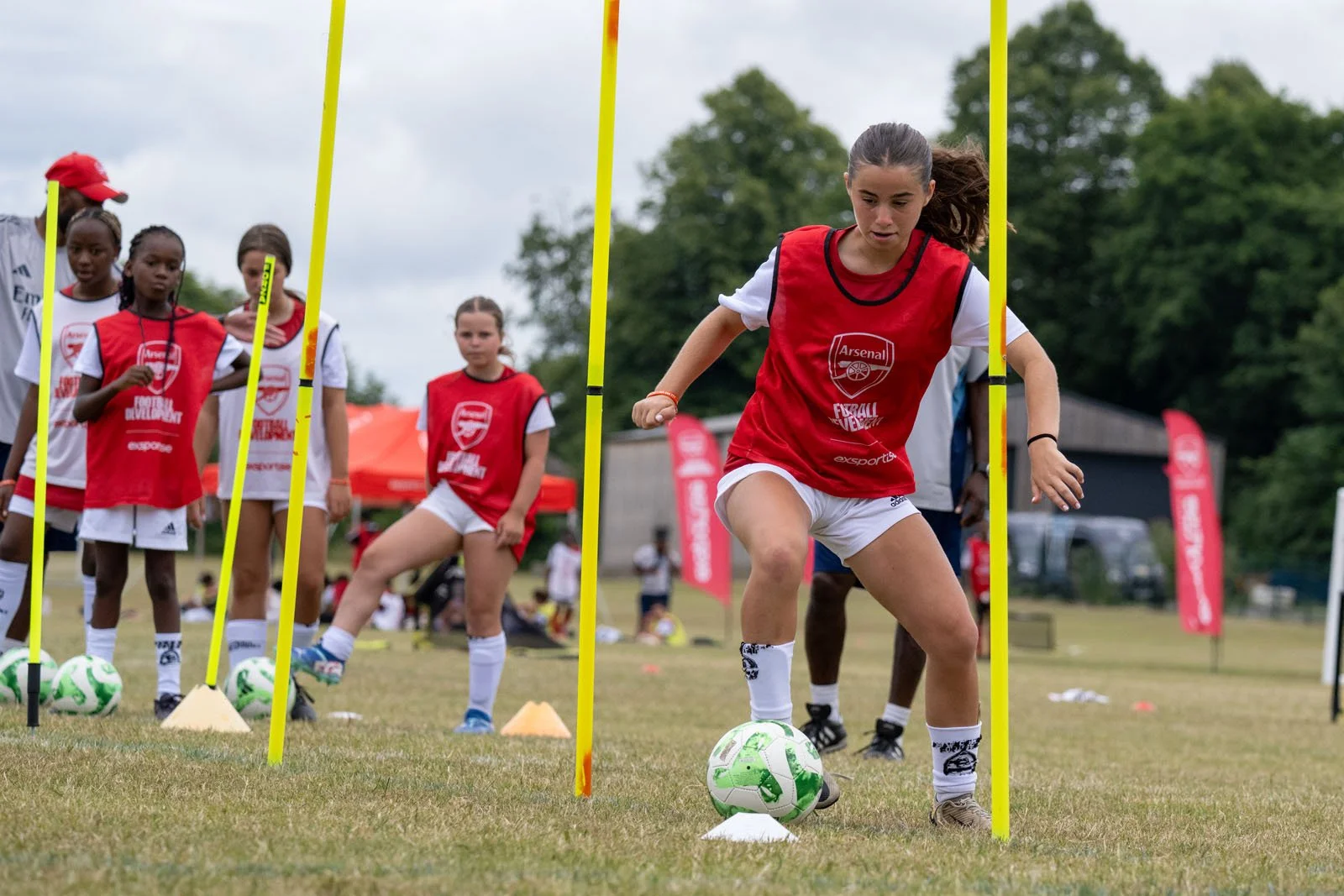 A soccer training session with young players and a coach on a grassy field, practicing dribbling around yellow training mannequins.