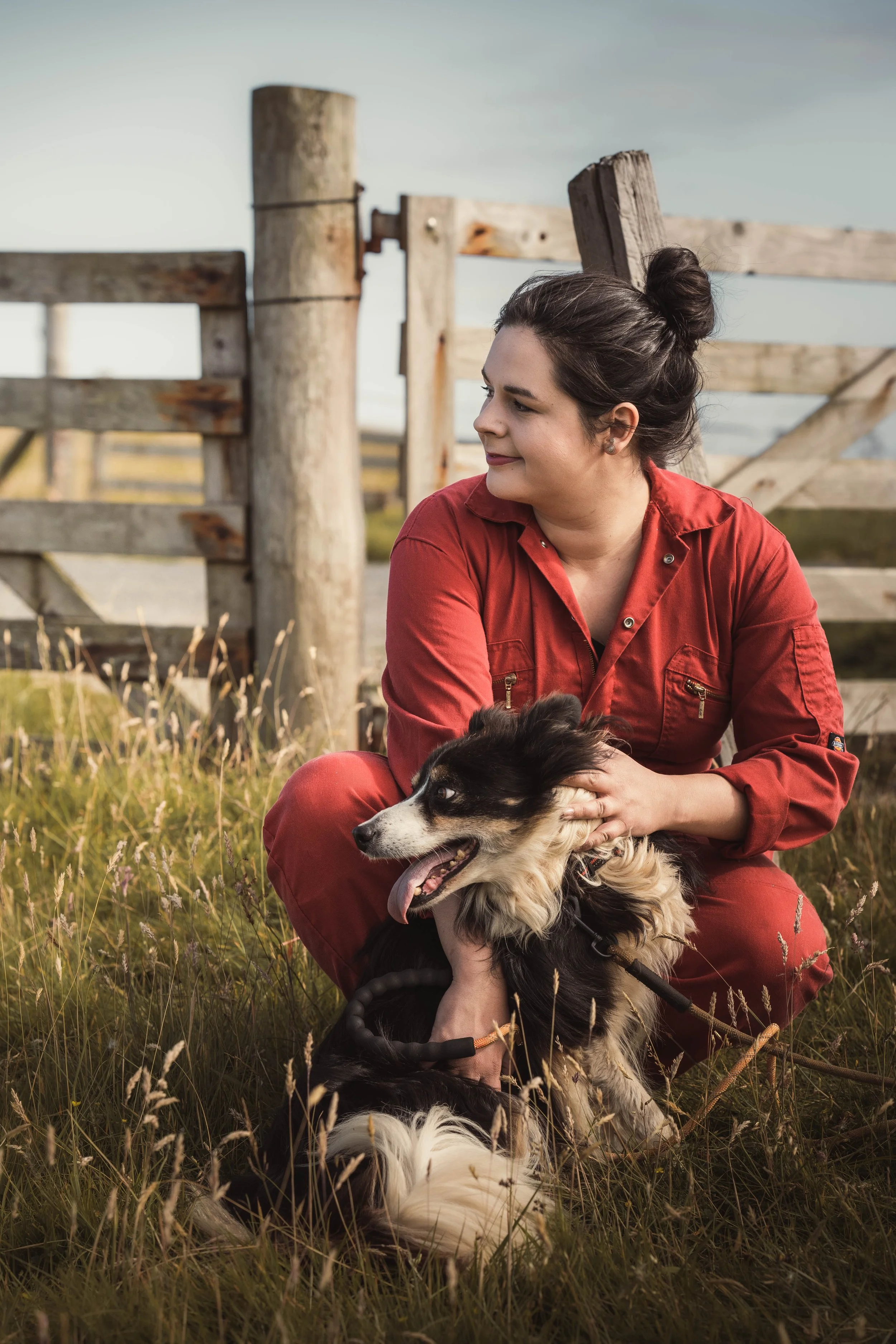 Marie a red jumpsuit sitting on grass, holding a black and white Australian Shepherd dog with its tongue out, near a wooden fence in an outdoor setting with tall grass and a partly cloudy sky.