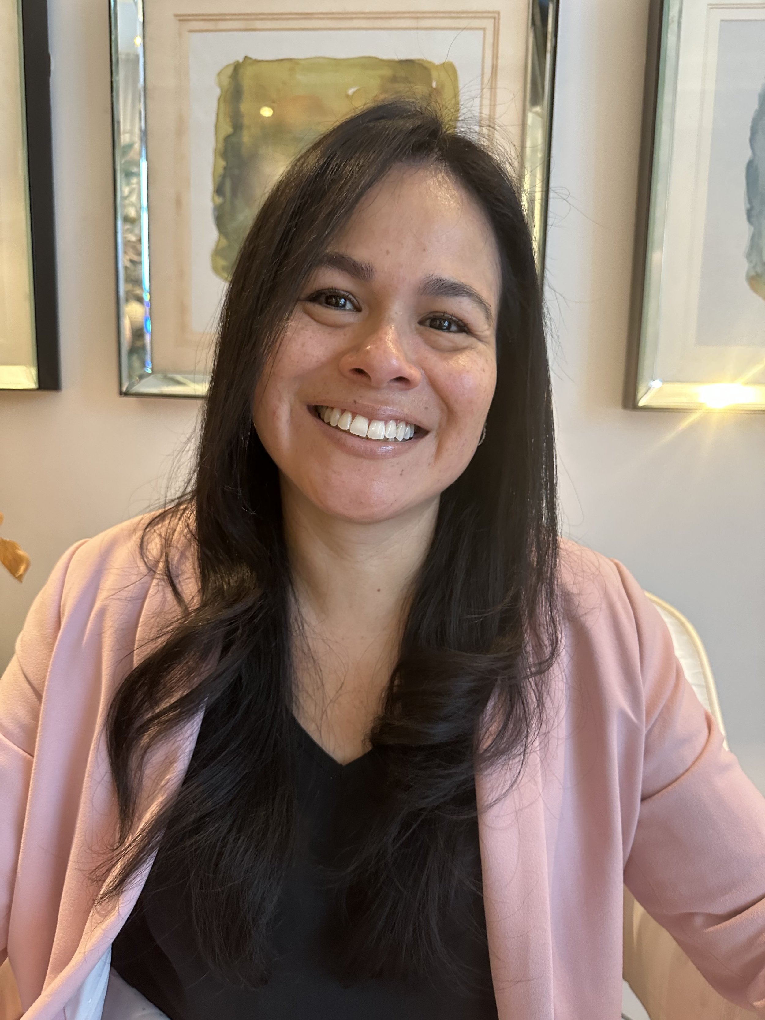 Smiling woman with long dark hair, wearing a pink blazer and black top, sitting indoors in front of framed artwork.
