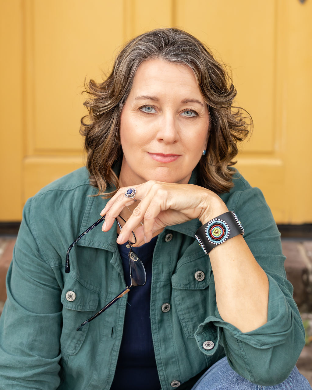 A woman with shoulder-length wavy brown hair and piercing blue eyes, wearing a green jacket, holding sunglasses, and displaying a colorful beaded bracelet on her wrist, sitting in front of a yellow background.