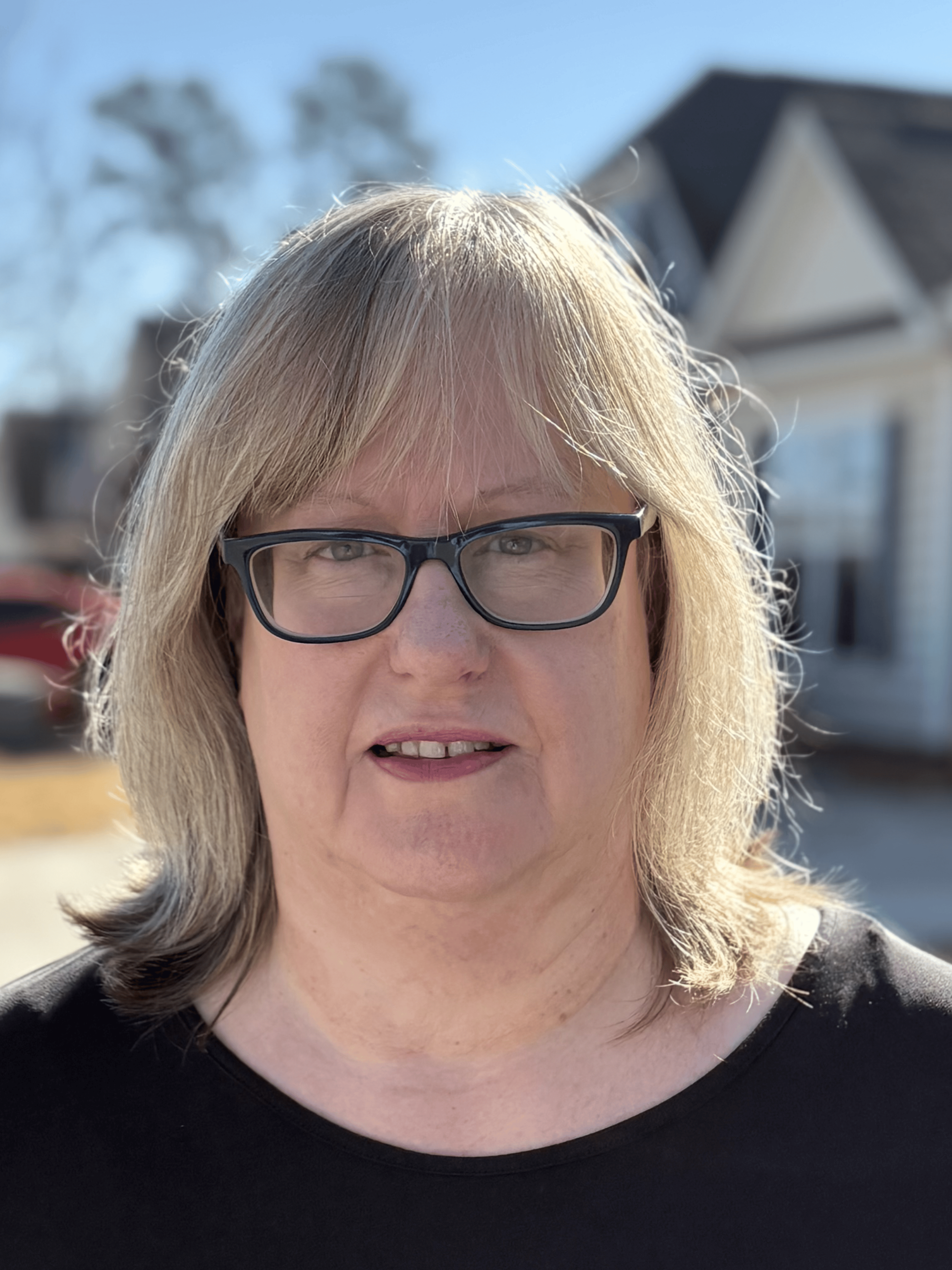 A woman with shoulder-length blonde hair, wearing black glasses and a black top, standing outdoors on a sunny day with houses and trees in the background.