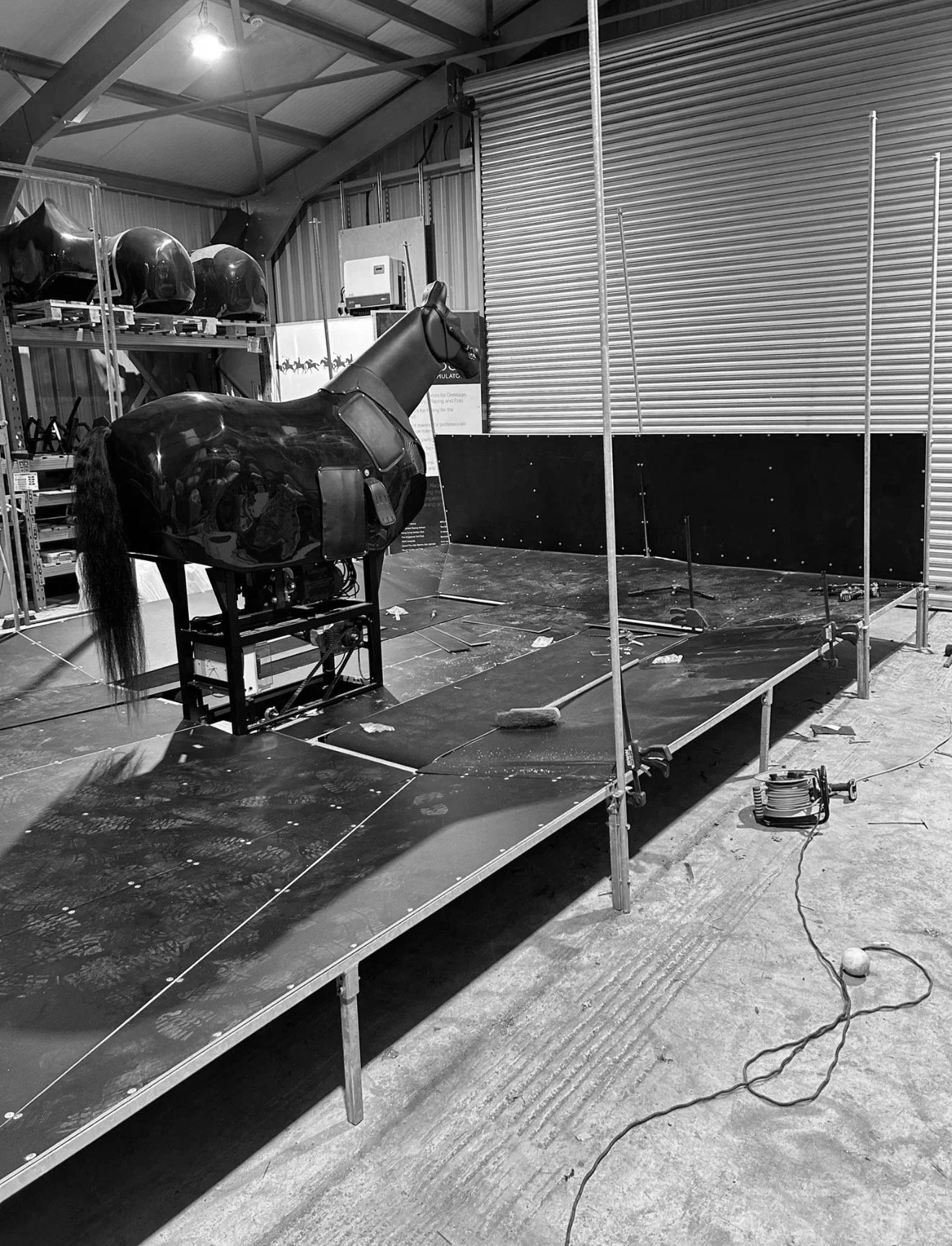 Black robotic horse on a platform in a warehouse or studio with equipment, helmets on shelves, and a black backdrop, suggesting a robotics or film set.