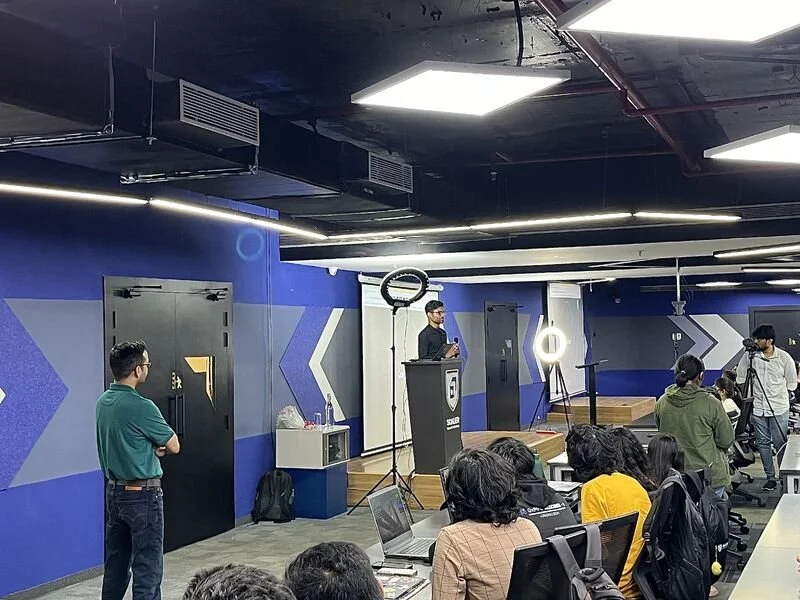 A man stands on a podium speaking to a group in a modern conference room with blue walls, LED lighting, camera equipment, and a seated audience.