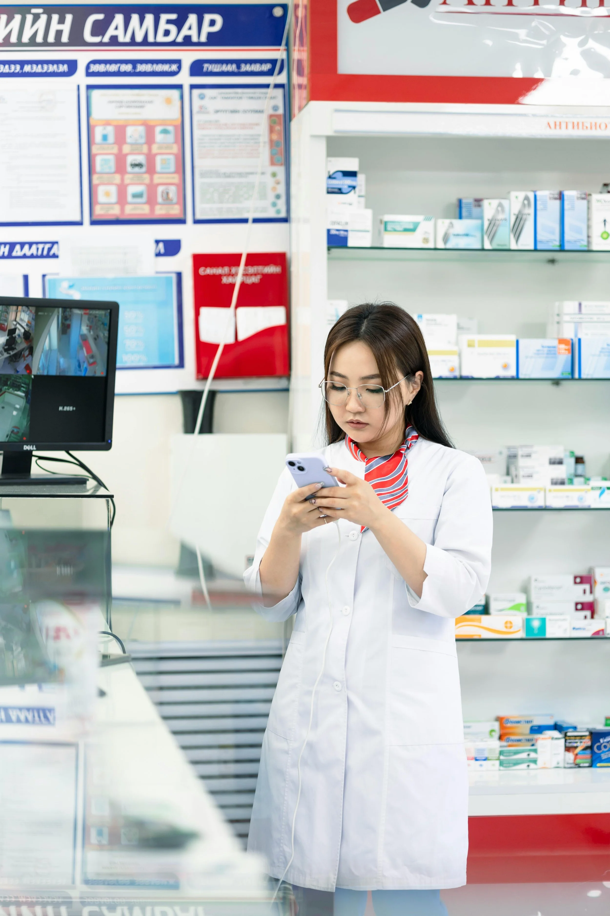 A young female pharmacist wearing glasses and a white lab coat, standing behind a pharmacy counter and looking at her smartphone, with shelves of medication and medical supplies in the background.