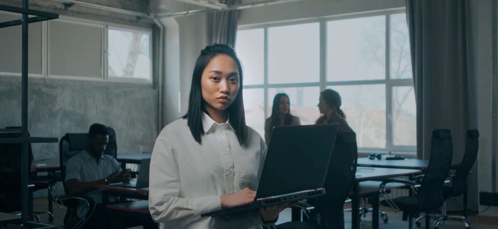 A woman in a white shirt holding a laptop in a modern office or classroom setting with large windows. In the background, three people are talking and another person is working on a laptop.