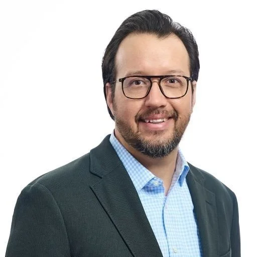 Professional man wearing glasses, a blue dress shirt, and a dark blazer, smiling against a plain white background.