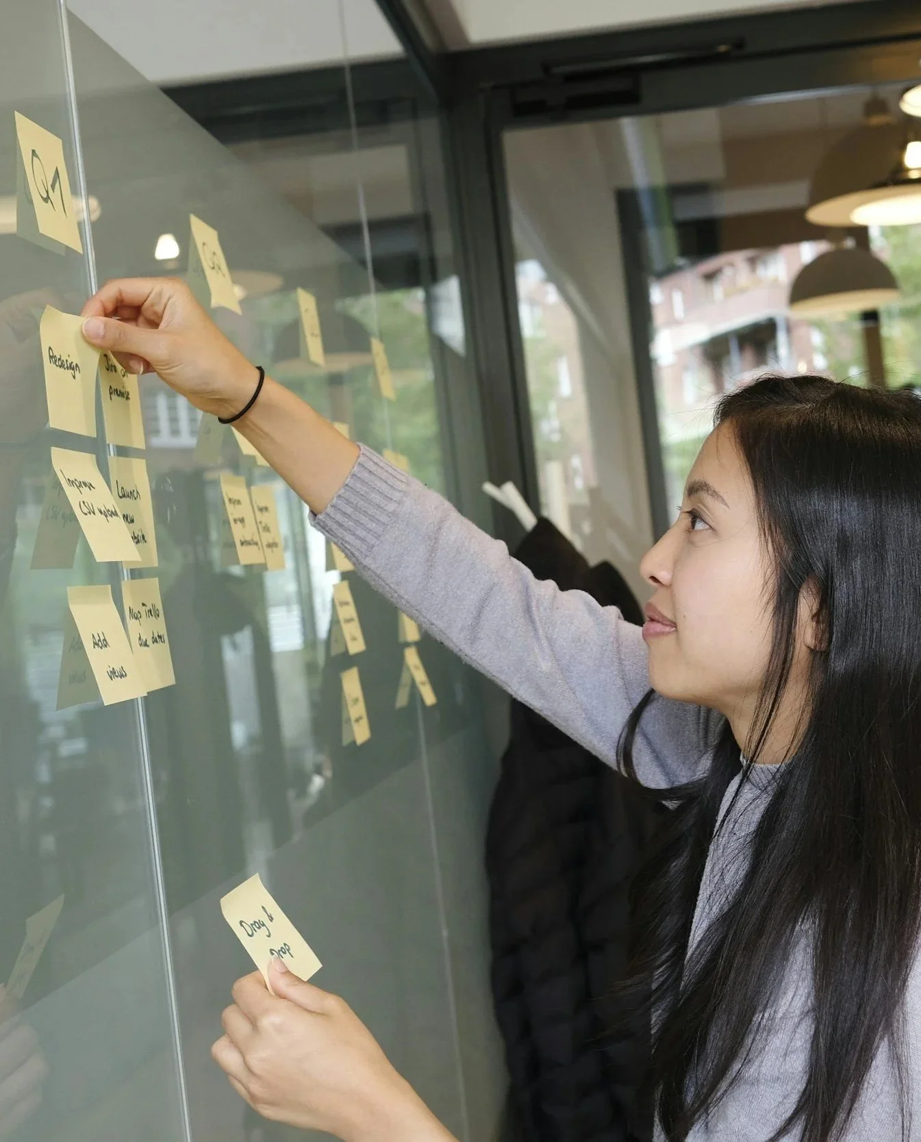 A woman with long black hair placing a sticky note on a glass wall covered with other sticky notes in a bright, modern office or coworking space.