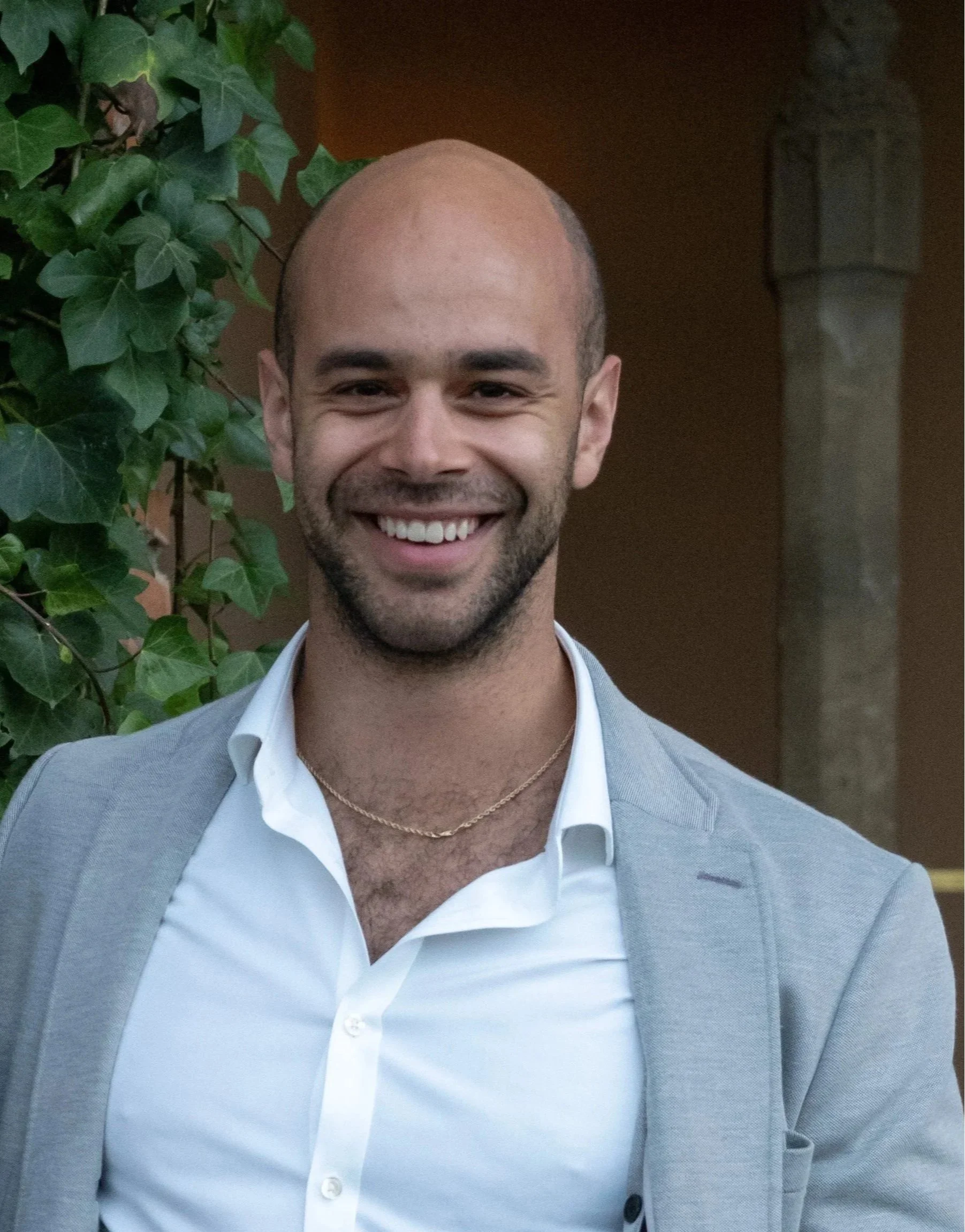 A smiling young man with a shaved head and beard, wearing a white dress shirt, light gray blazer, and a gold chain, standing outdoors in front of green ivy and a brown wall.