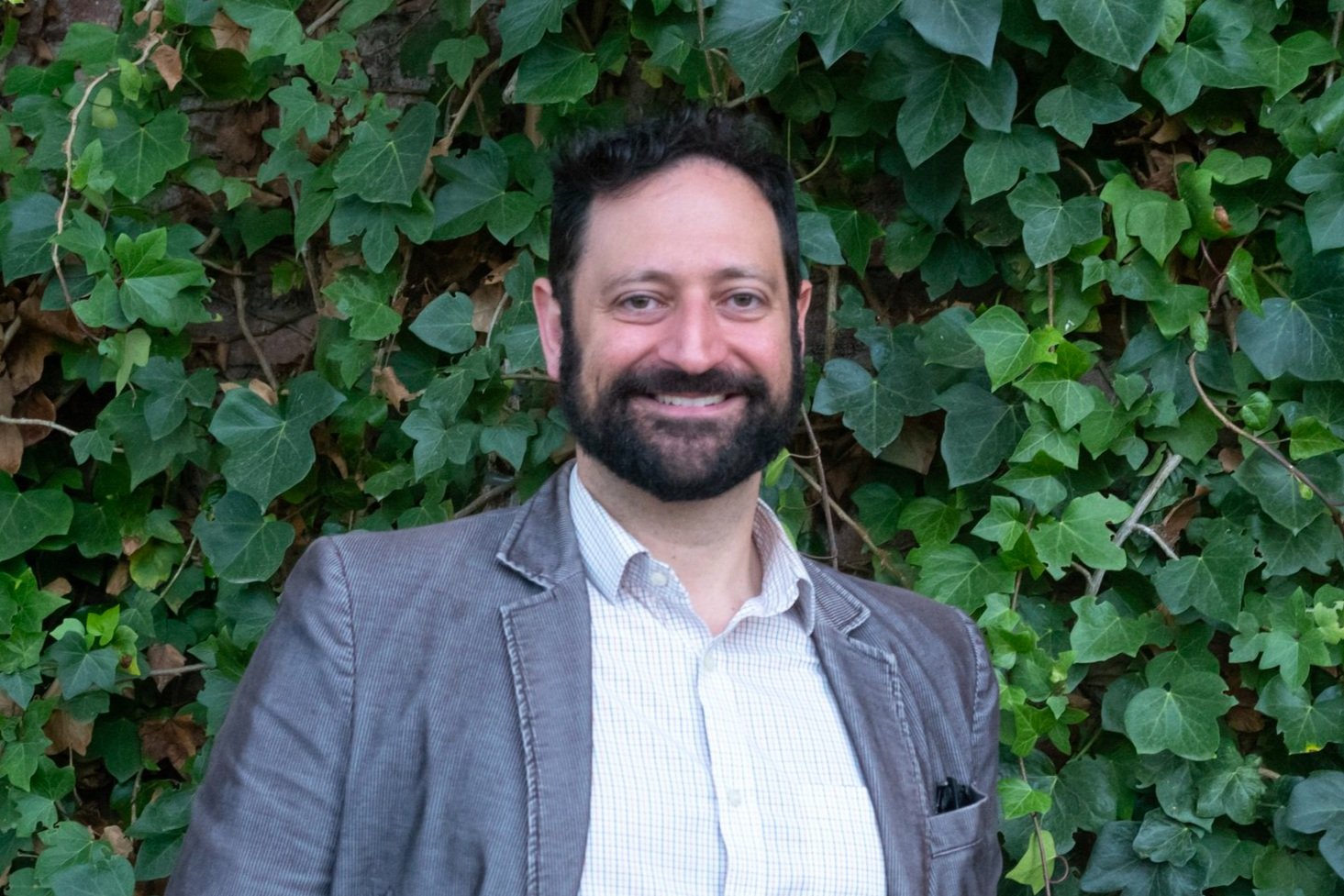 A man with dark hair and a beard, smiling, standing in front of a background of green ivy leaves. He is wearing a light-colored checkered shirt and a gray blazer.